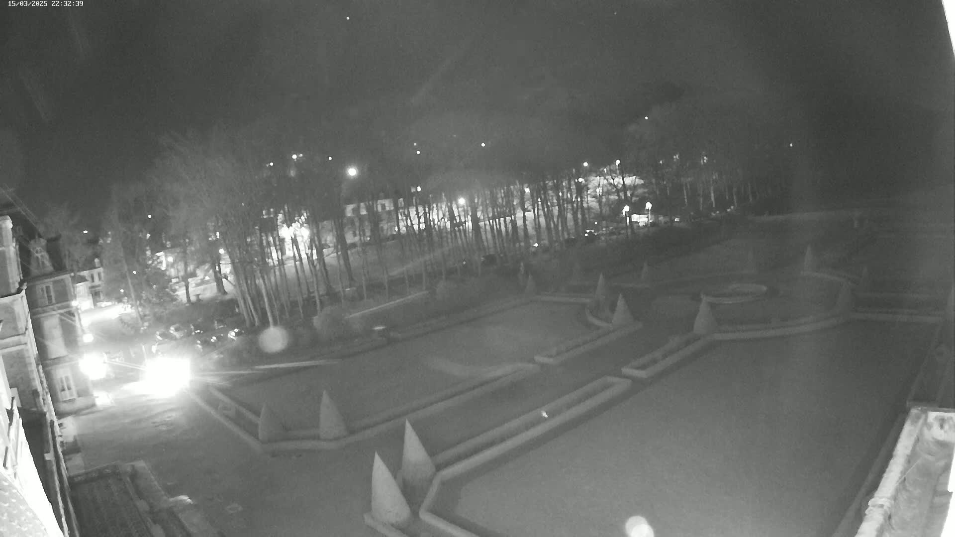 A nighttime, low-light view shows a formal garden with geometric patterns and a building in the background, lit by several streetlights.