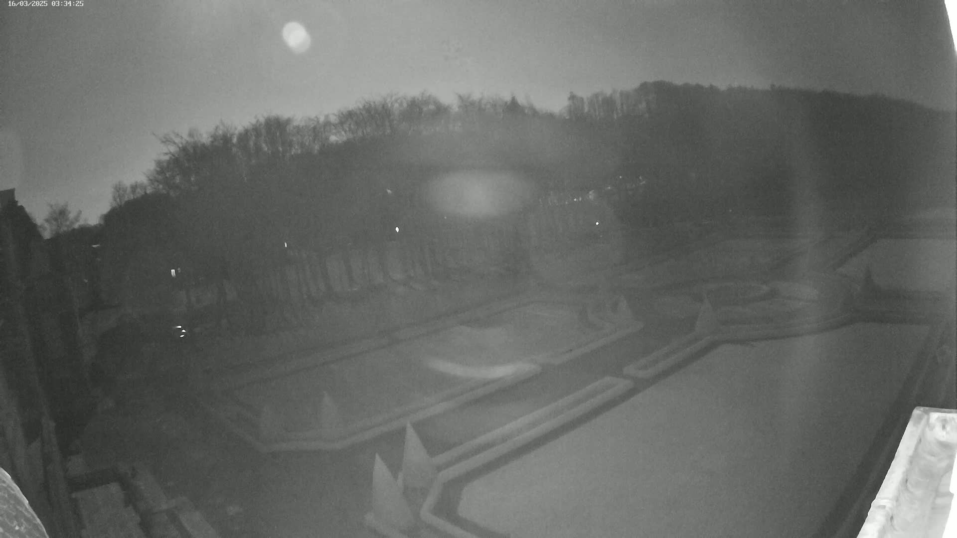 A nighttime, low-light view shows a dark, expansive garden with geometrically patterned pathways and pools, bordered by a line of trees under a mostly clear sky.