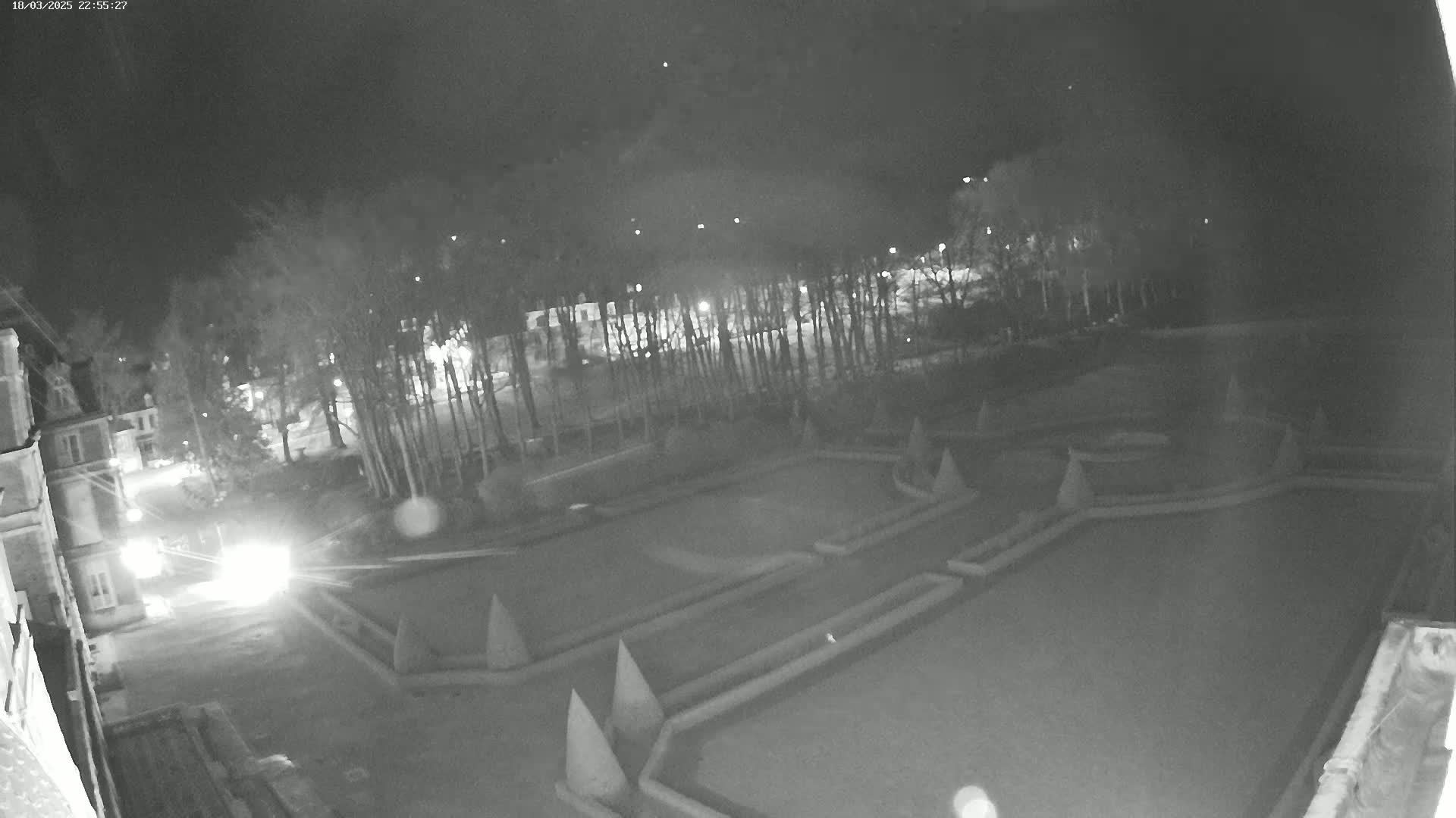 A nighttime, black-and-white view of an expansive, geometrically-designed garden bordered by trees and a building, illuminated by faint light sources.