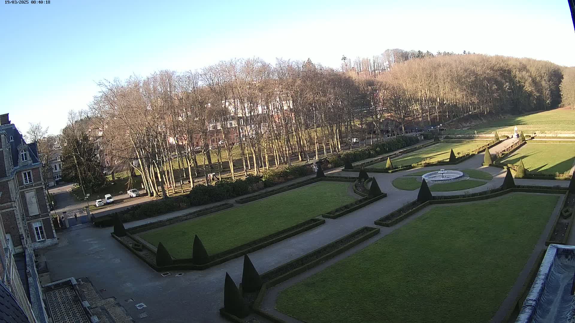 A formal garden with a fountain is visible from an elevated viewpoint, on a sunny day with leafless trees in the background.