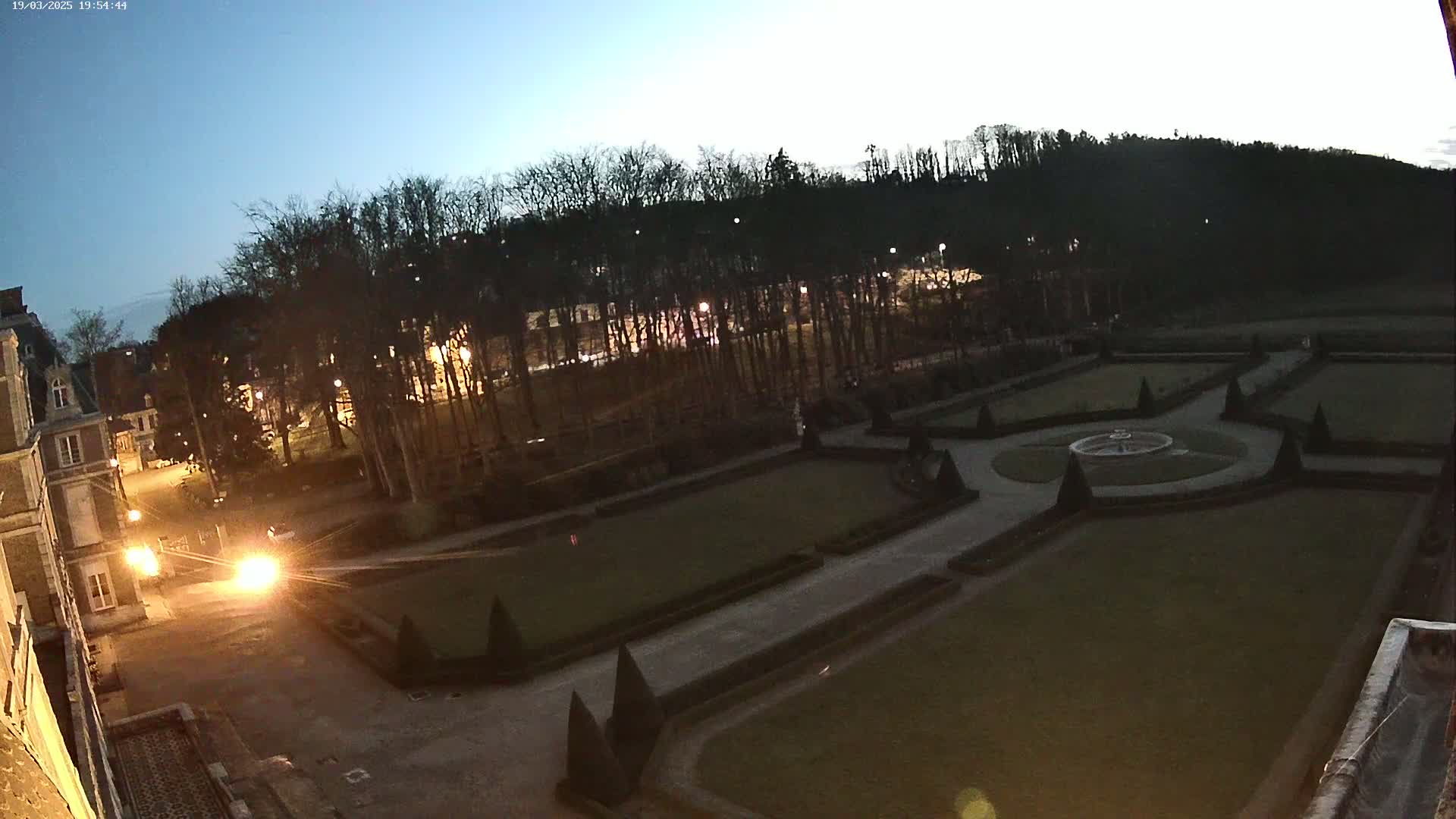 An expansive, formal garden with geometrically shaped lawns and hedges is visible at twilight, viewed from an elevated position near a large building.