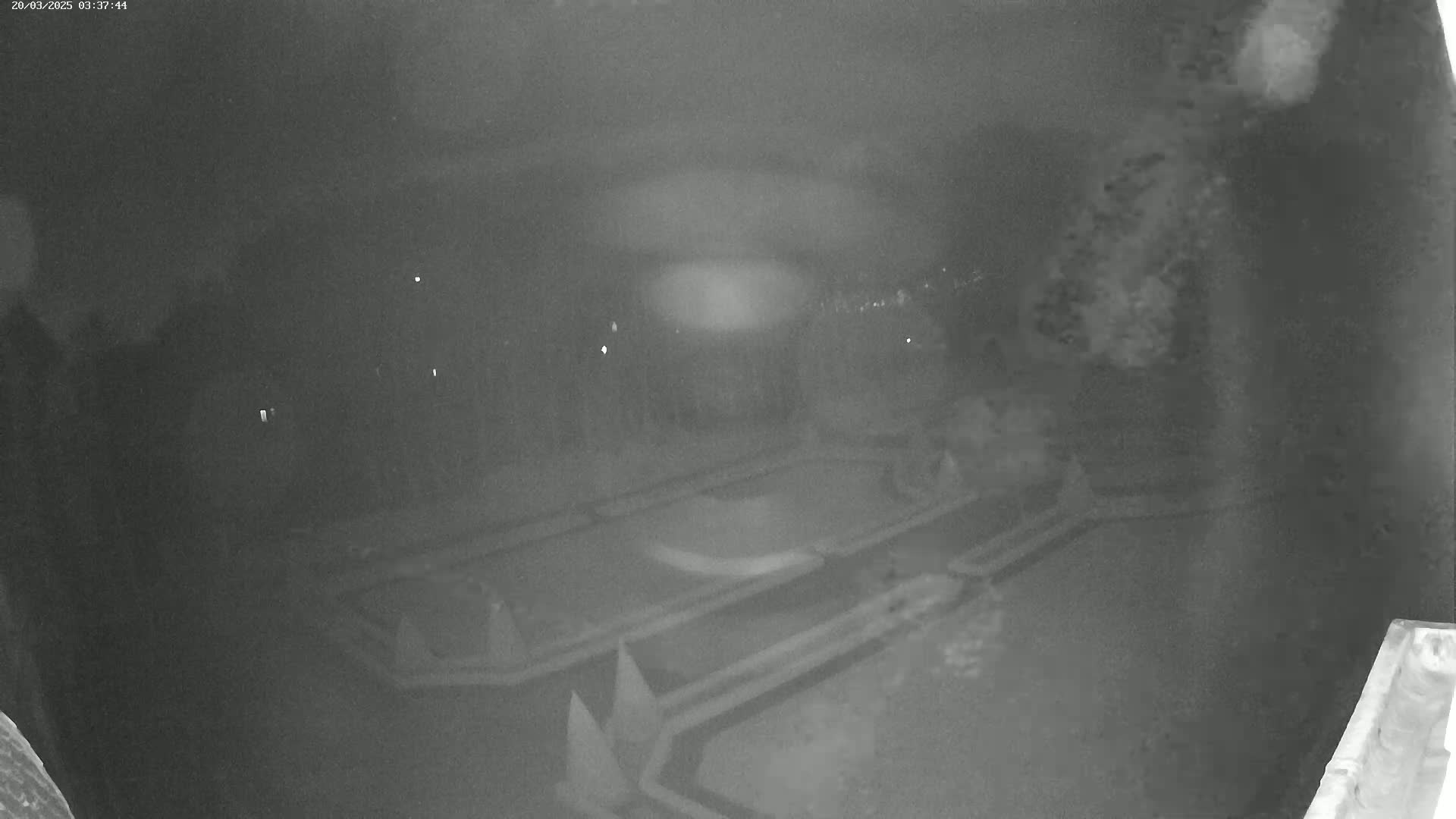 A nighttime, low-light image shows an aerial view of a dark, geometrically patterned area, possibly a park or plaza, with some blurry lights visible in the distance.