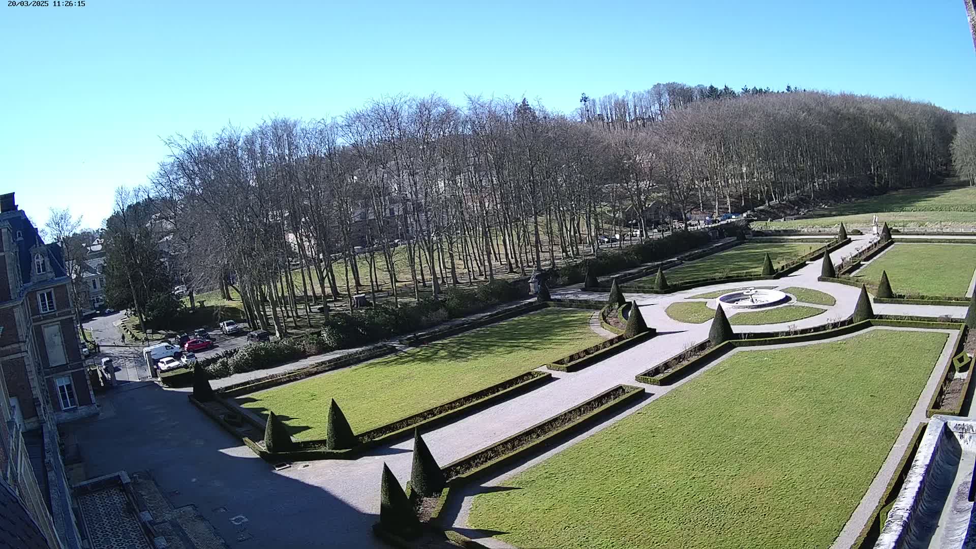 A formal garden with a central fountain, manicured lawns, and neatly trimmed hedges is situated next to a large building on a sunny day.