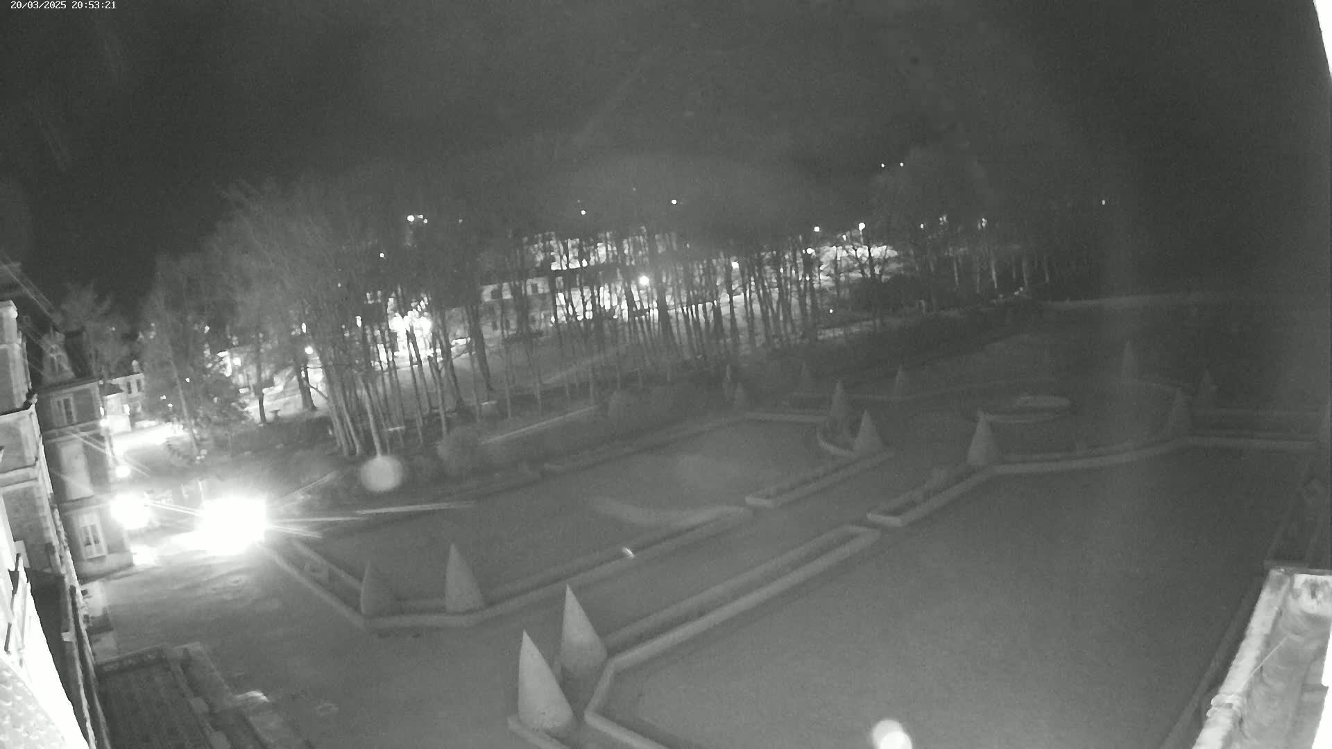 A nighttime, low-light view of an ornate garden with hedges and conical topiary, situated near a building and a road with a vehicle's headlights visible.