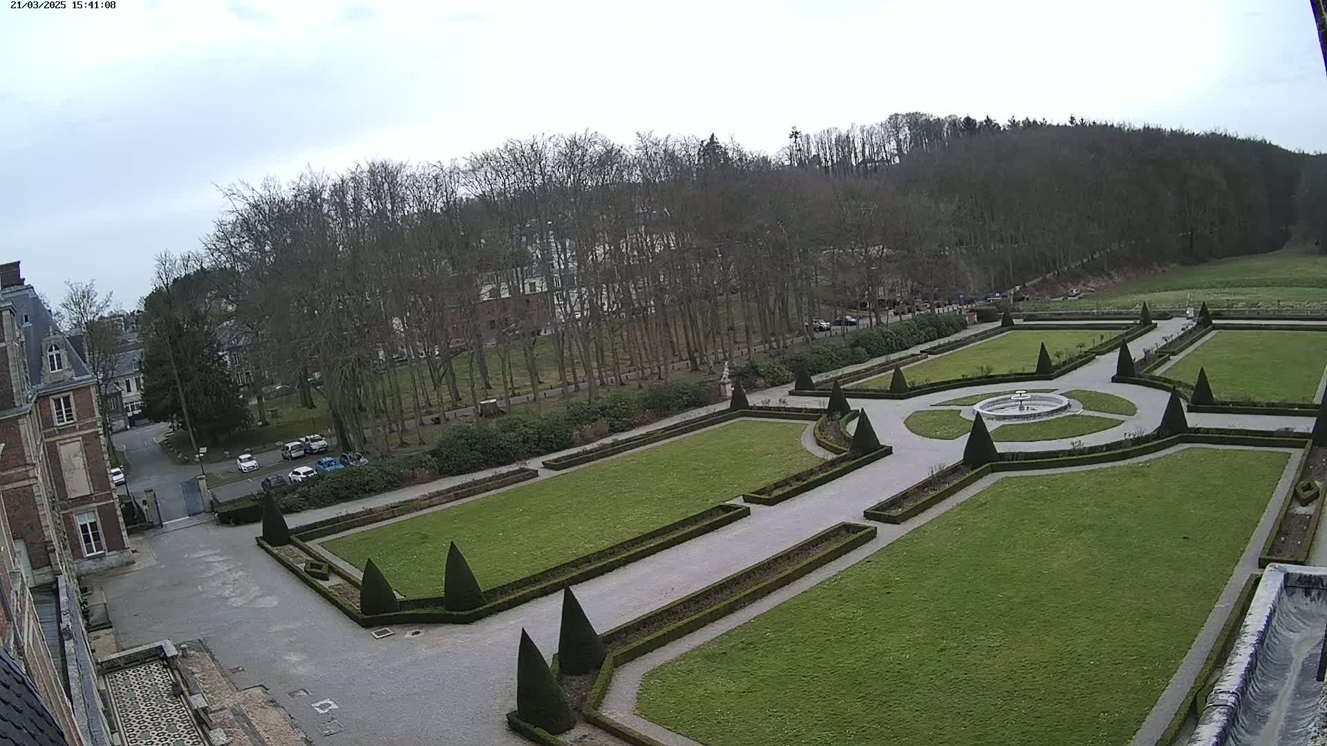 An expansive formal garden with manicured lawns, hedges, and a central fountain is situated beside a large building, all under a cloudy sky.