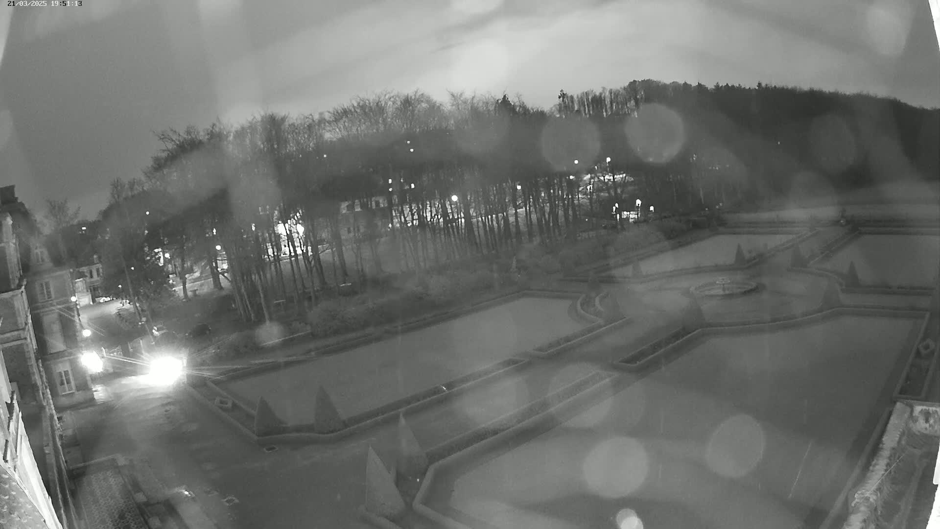 A nighttime, rain-speckled view of an ornate formal garden with a central fountain, situated beside a building and a tree line under a cloudy sky.