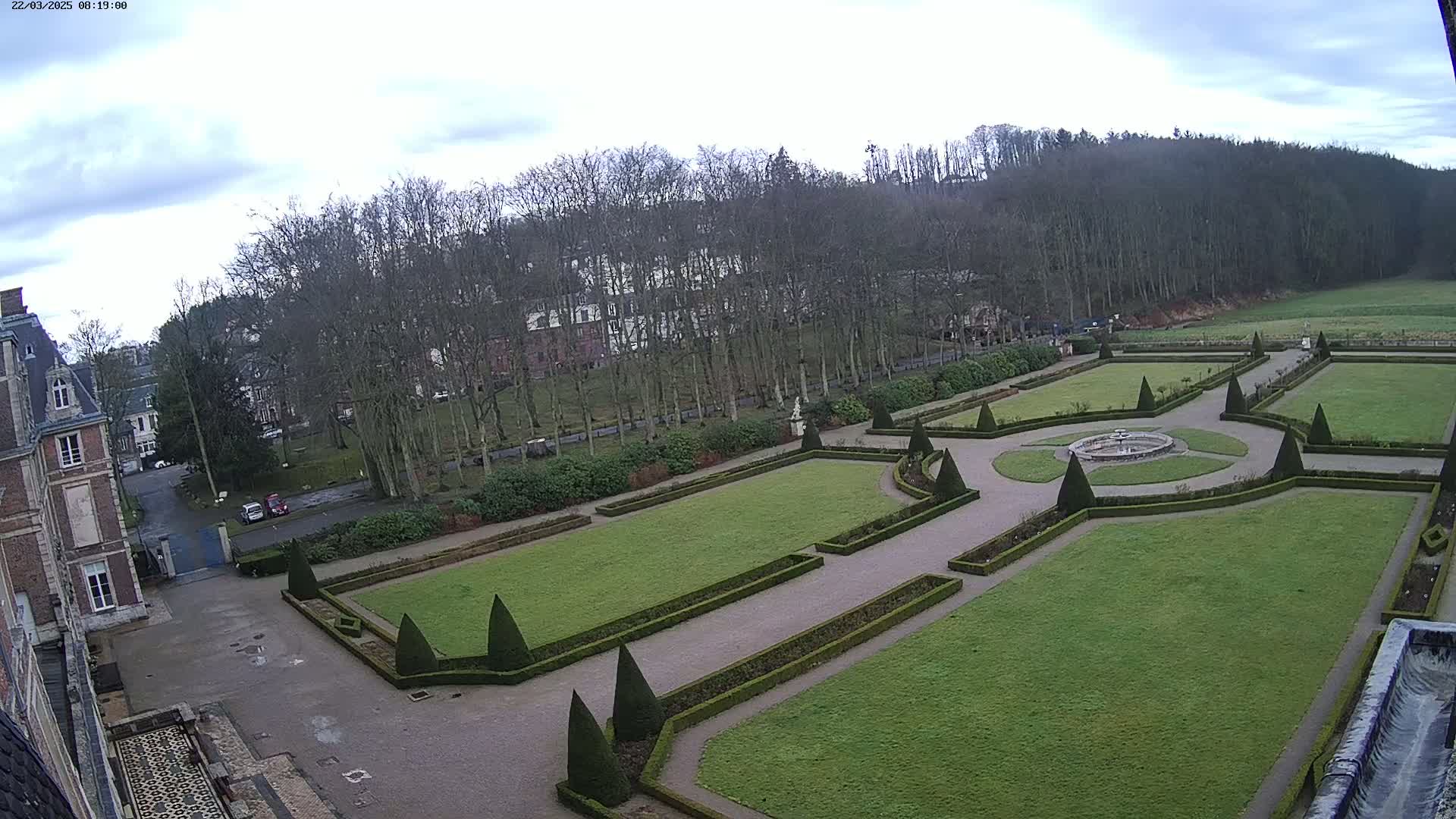A formal garden with manicured lawns, hedges, and a central fountain is situated in front of a brick building under a cloudy sky.