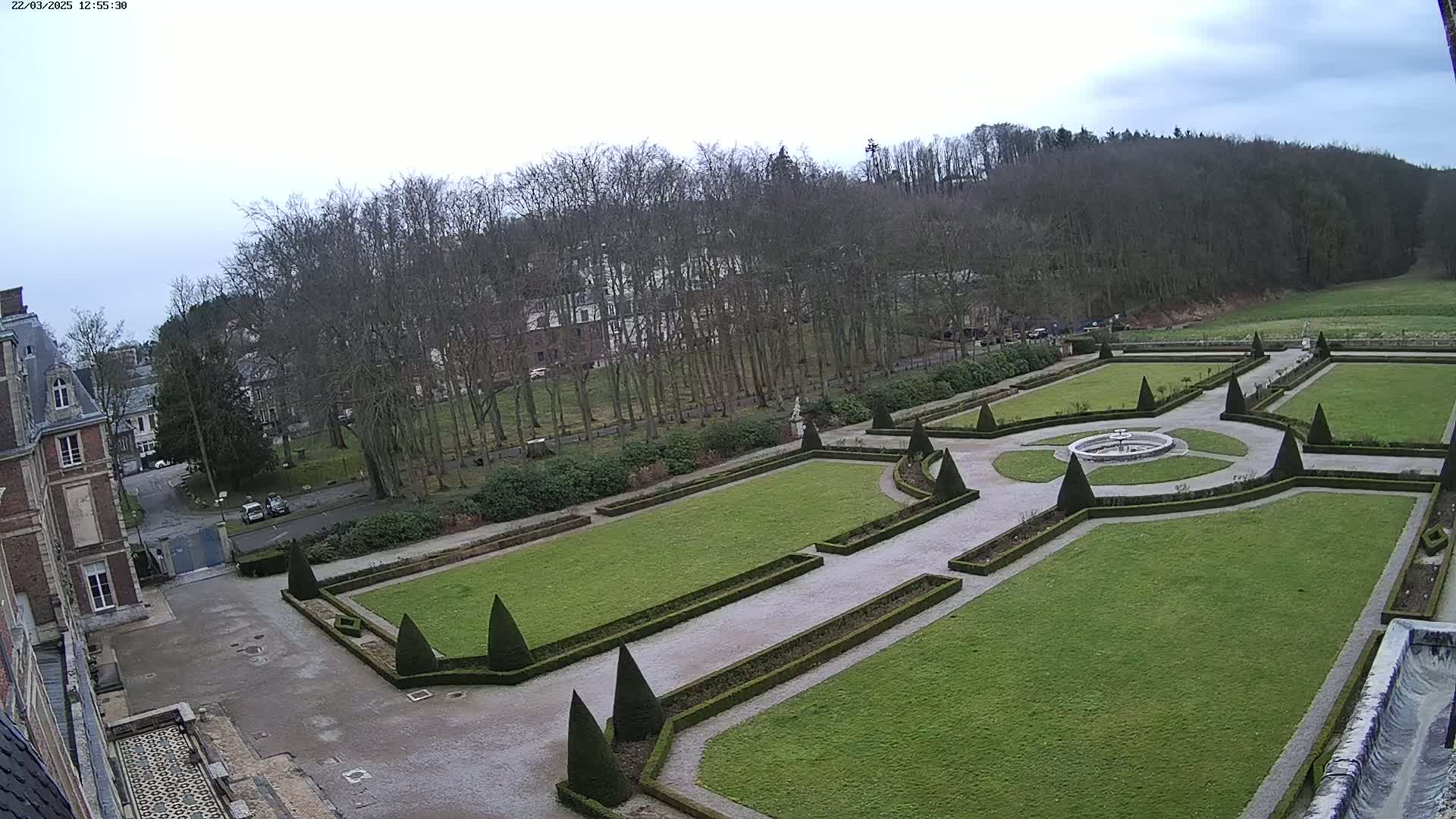 An ornate, geometric garden with a central fountain is viewed from above, under an overcast sky, adjacent to a large brick building and a wooded area.