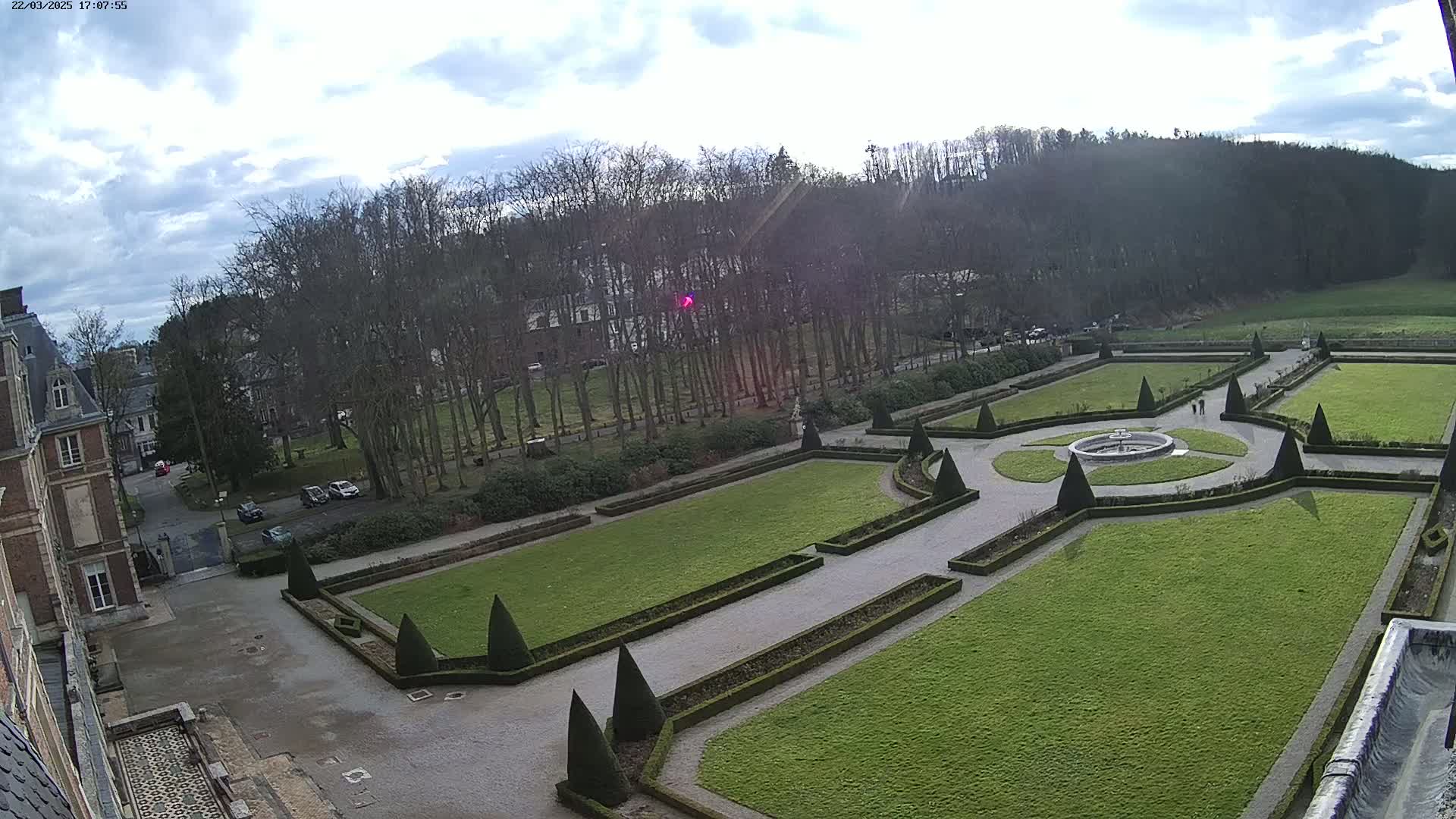 A formal garden with geometrically shaped lawns and hedges, a central fountain, and a chateau in the background under a partly cloudy sky.