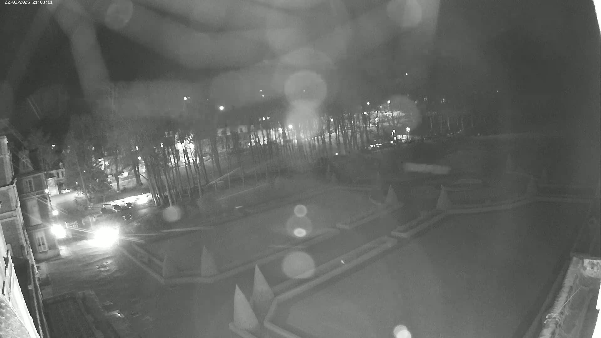 A nighttime, rainy view of a courtyard with geometric landscaping features and a road with cars visible in the distance.