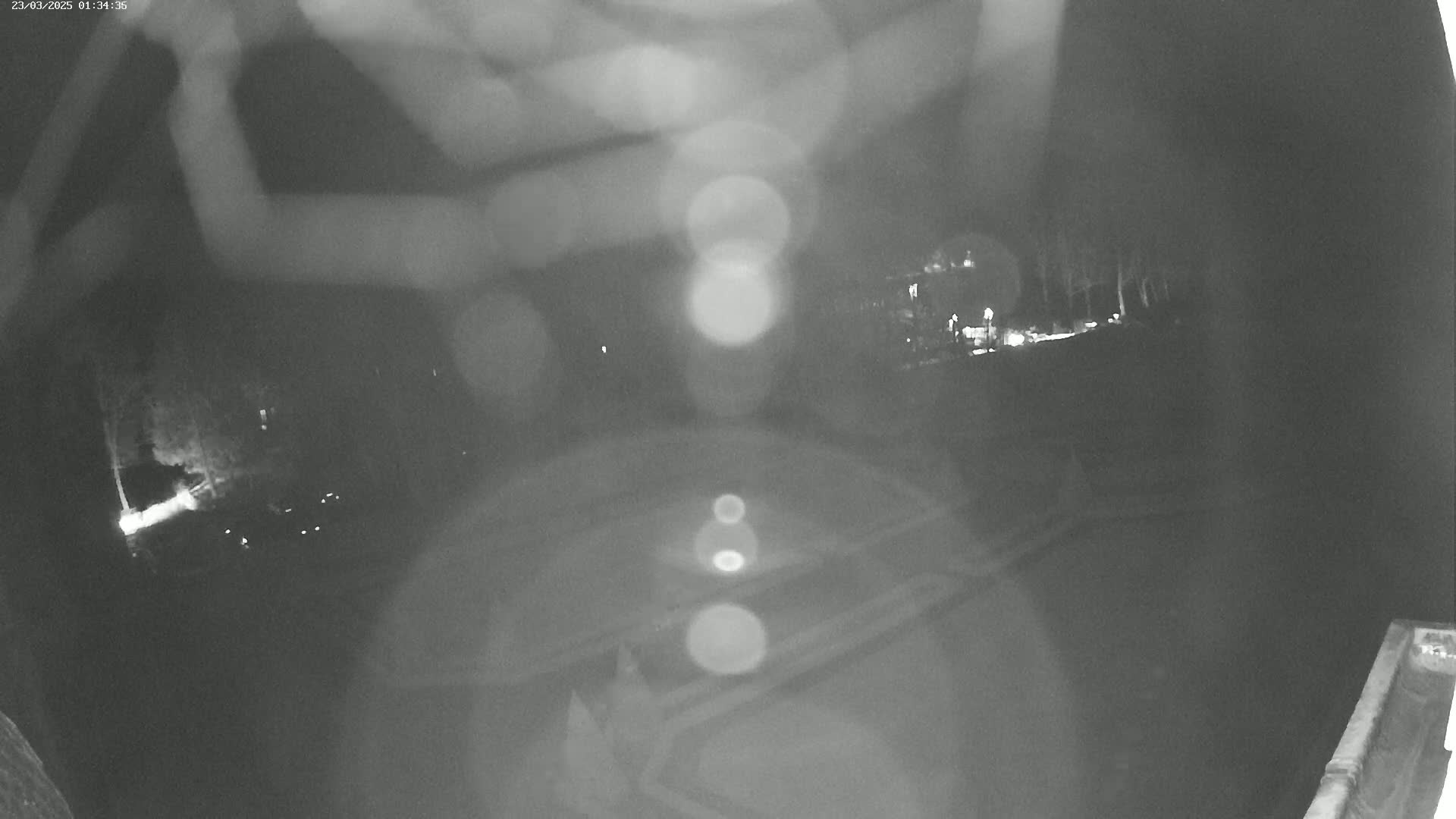 A nighttime, blurry, outdoor security camera view shows a dark area with some lights and indistinct shapes in the distance.