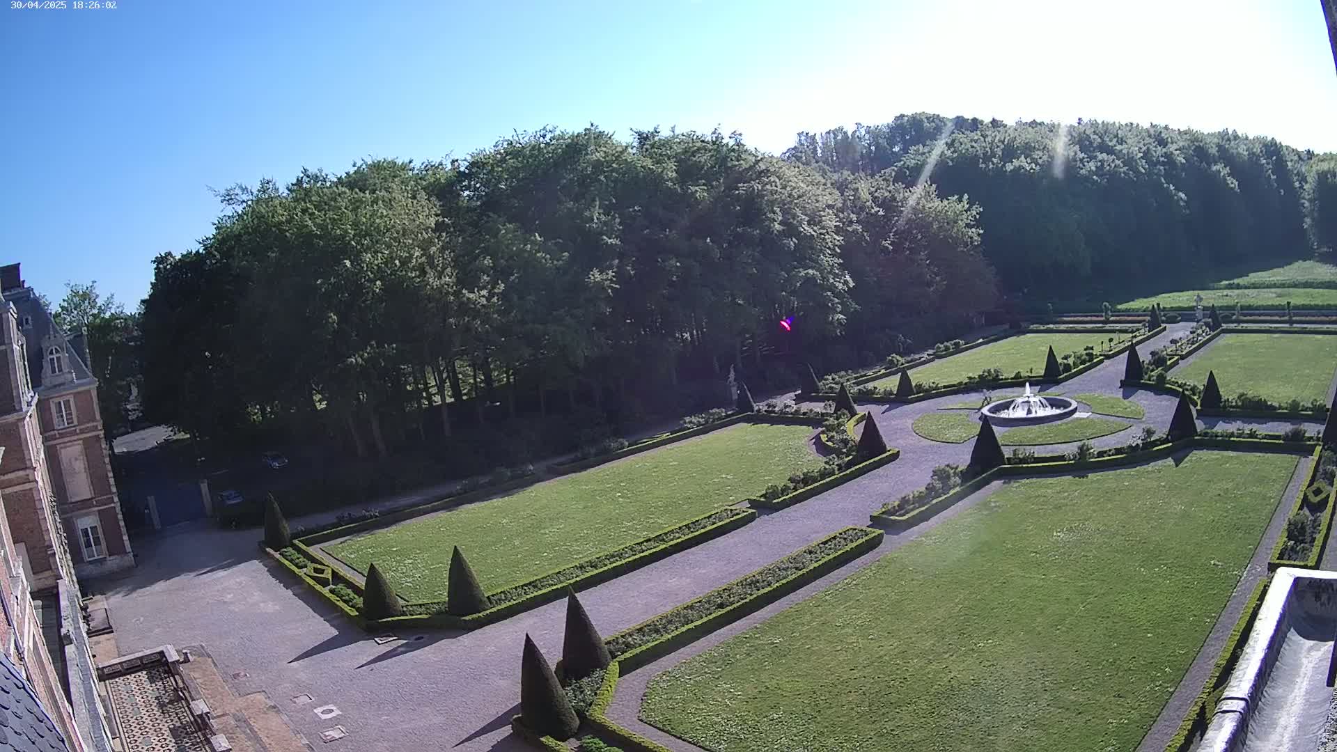 A high-angle view shows a formal garden with manicured lawns, hedges, and a central fountain, bordered by a large stone building and a line of trees under a sunny sky.