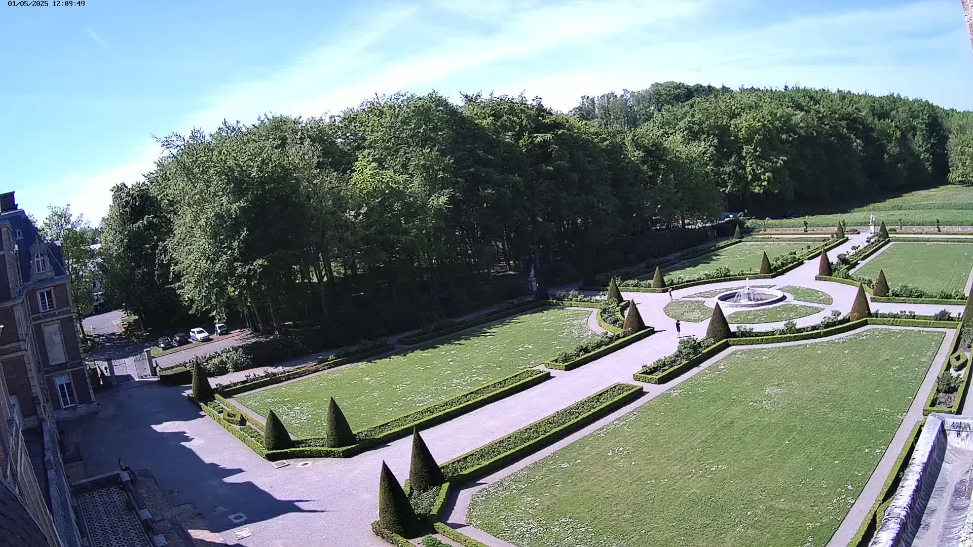A formal garden with a fountain is situated in front of a large building, surrounded by a lush green forest under a clear, sunny sky.