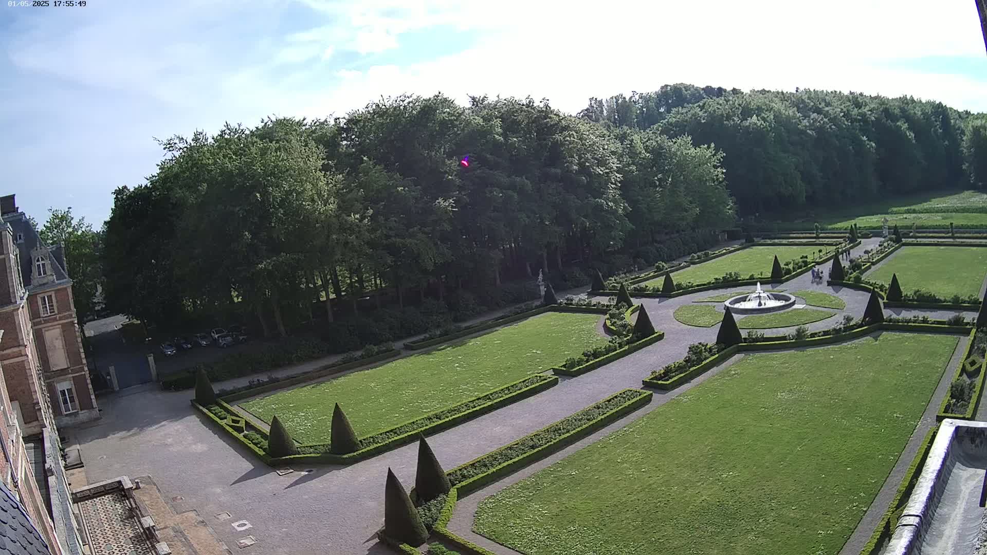 An elevated view of a formal garden with a fountain, manicured hedges, and a large expanse of lawn, bordered by a forest under a partly sunny sky.