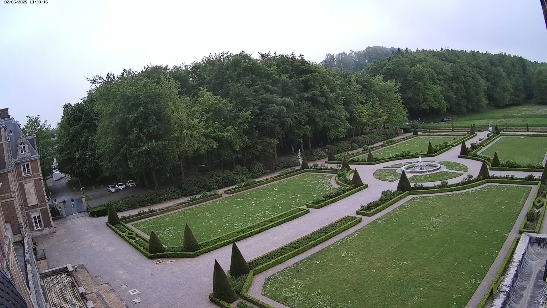 An overcast day reveals a formal garden with manicured lawns, hedges, and a central fountain, adjacent to a large brick building and a surrounding forest.