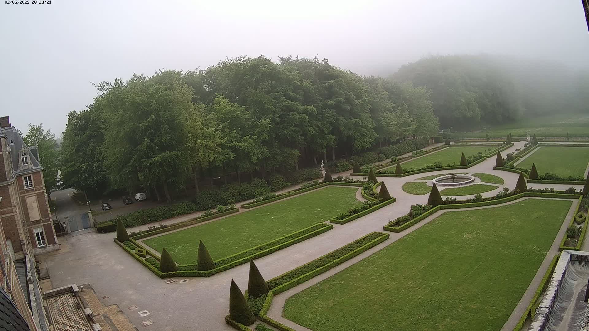 A formal garden with manicured hedges and a central fountain is situated behind a large building on a foggy day.