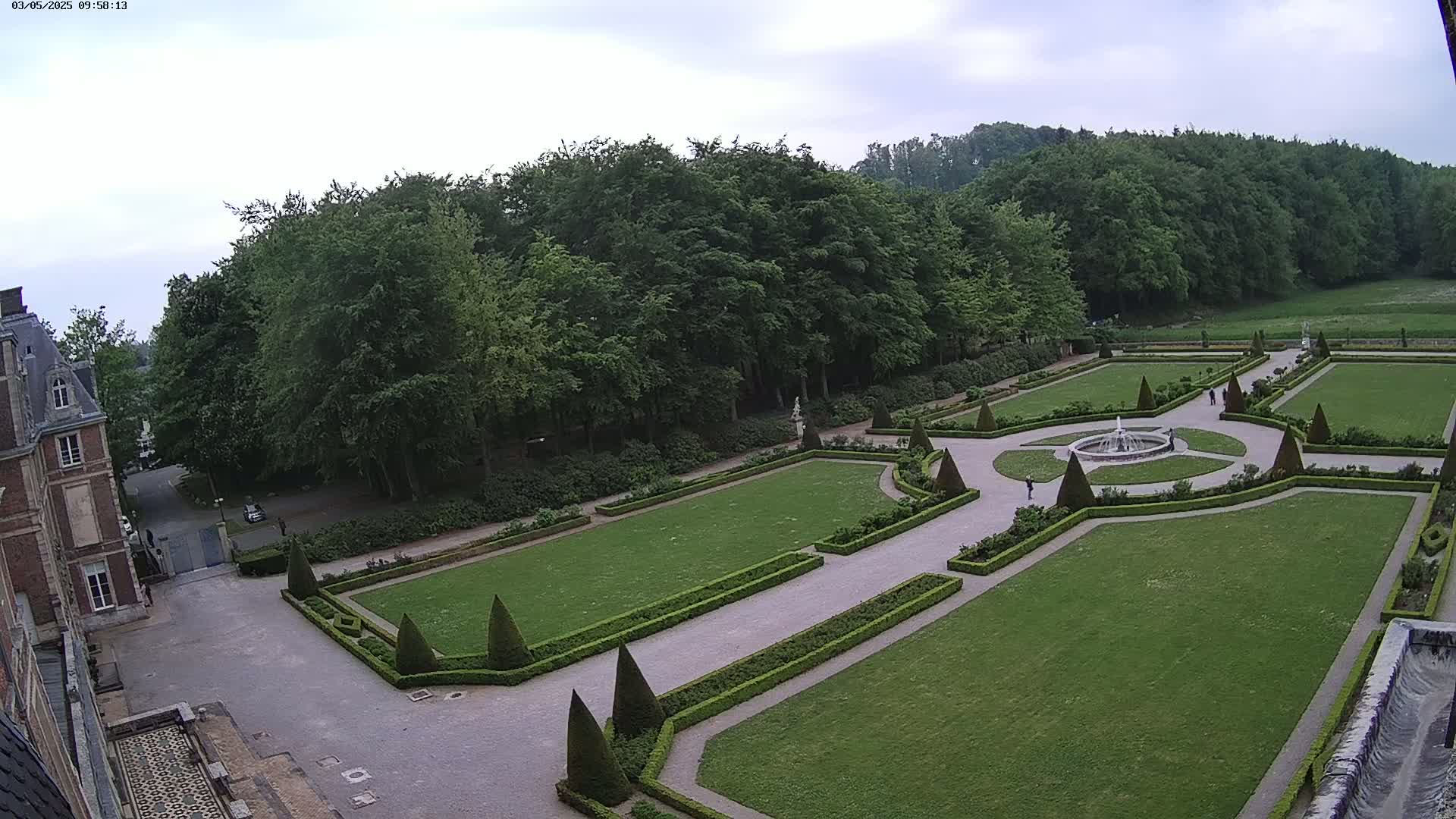 An expansive formal garden with a central fountain is visible from an elevated viewpoint, under an overcast sky, next to a large brick building and a surrounding forest.