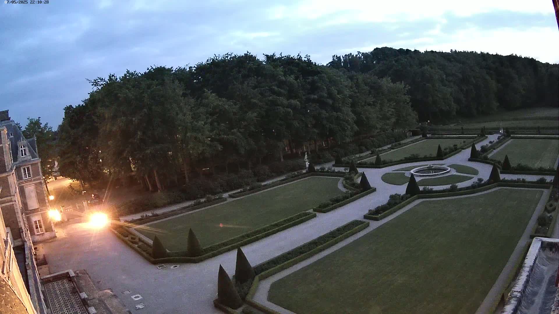 An expansive, formal garden with manicured lawns, hedges, and a central fountain is viewed from above at dusk, next to a large building.