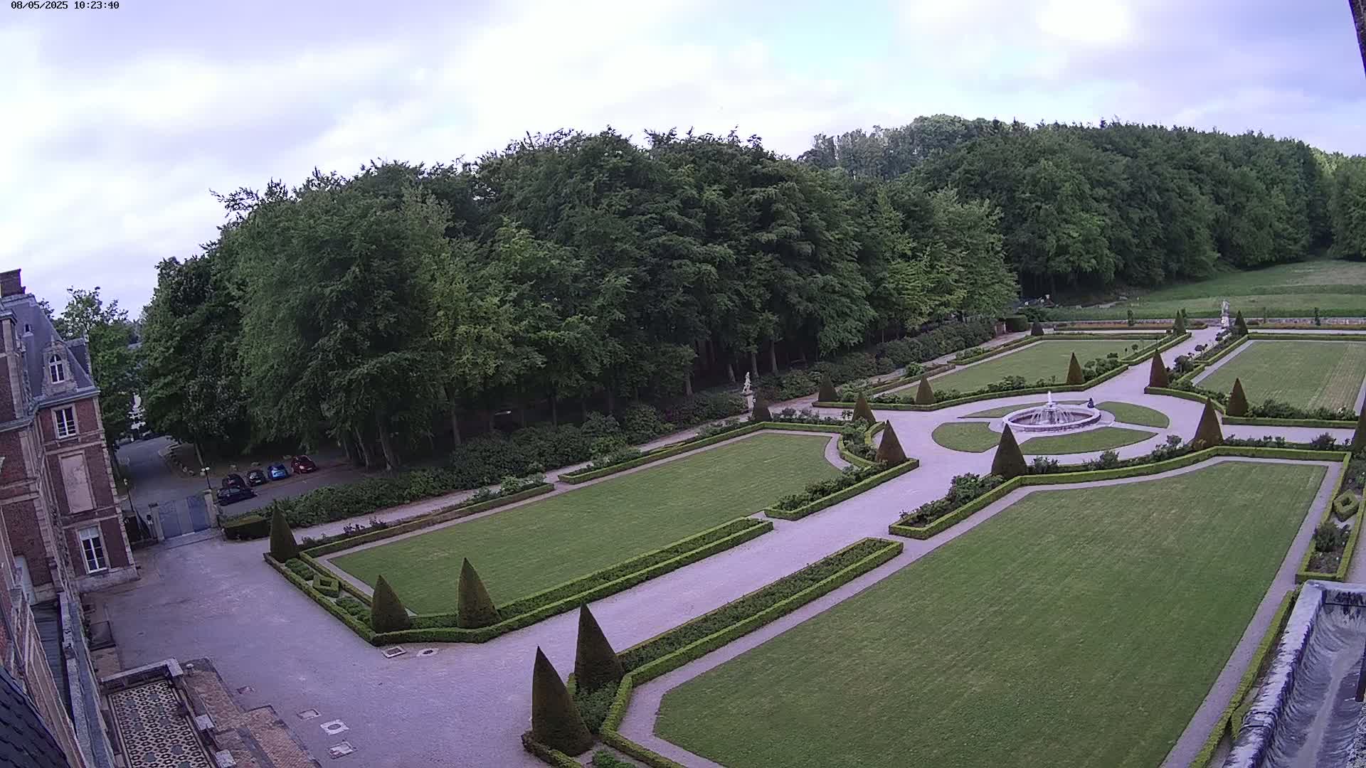 An aerial view of a formal garden with a fountain at its center, bordered by manicured hedges and lawns, and a building visible in the background under a partly cloudy sky.
