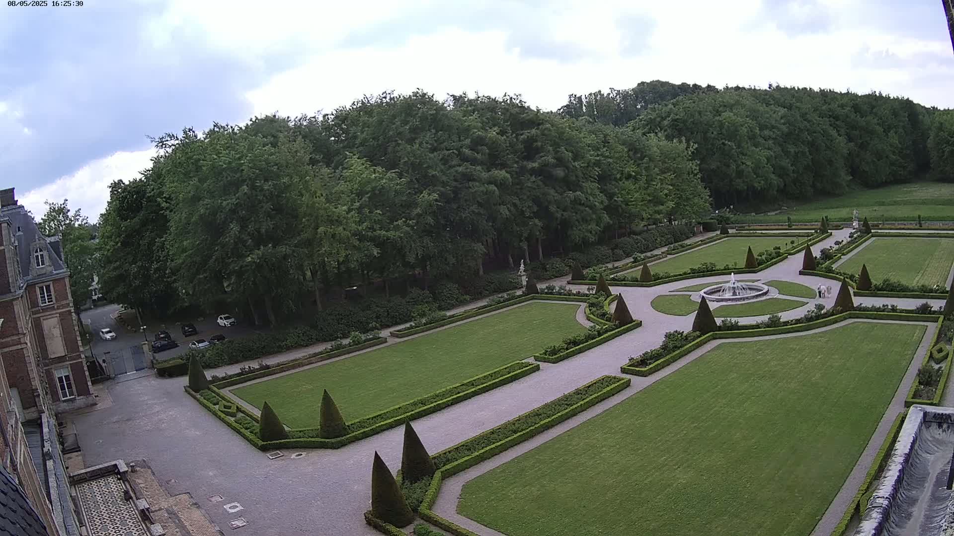 An expansive formal garden with a central fountain, manicured lawns, and hedges, is adjacent to a large building under a partly cloudy sky.