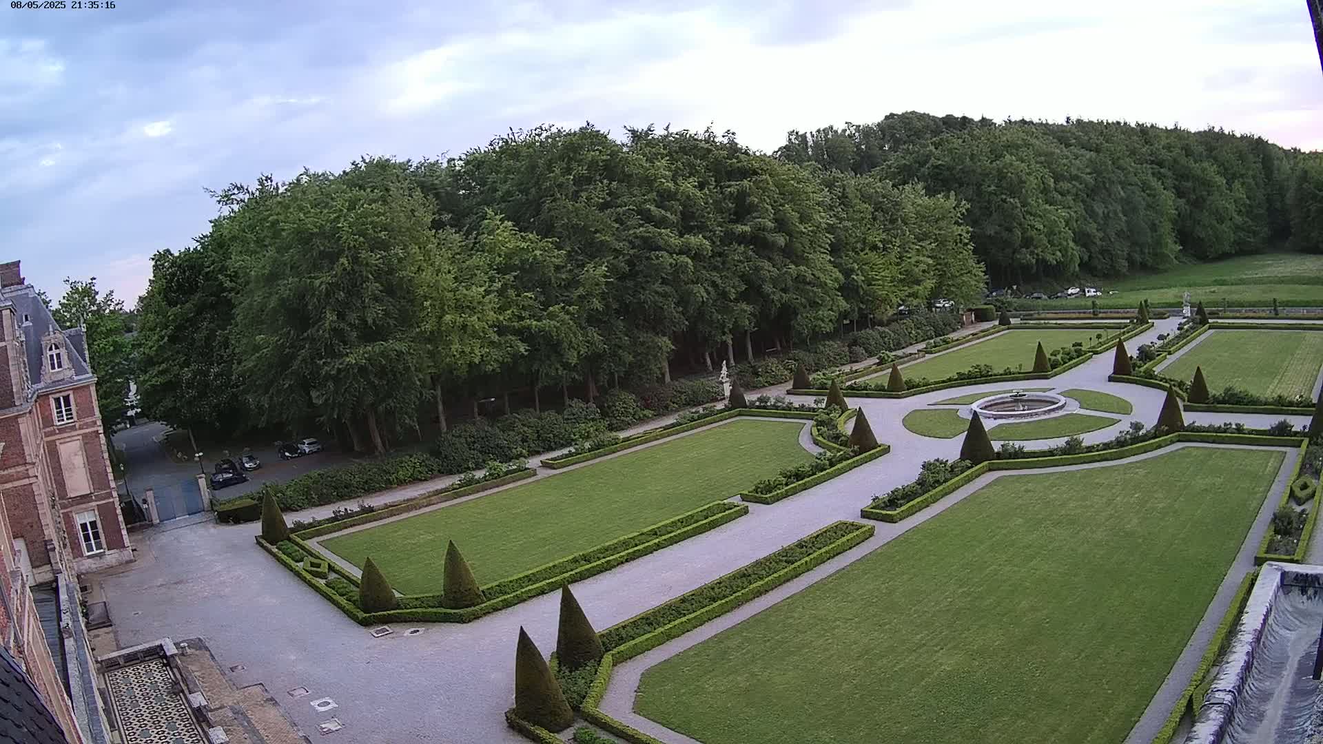 An aerial view shows a formal garden with manicured lawns, hedges, and a central fountain, adjacent to a large brick building under a partly cloudy sky.