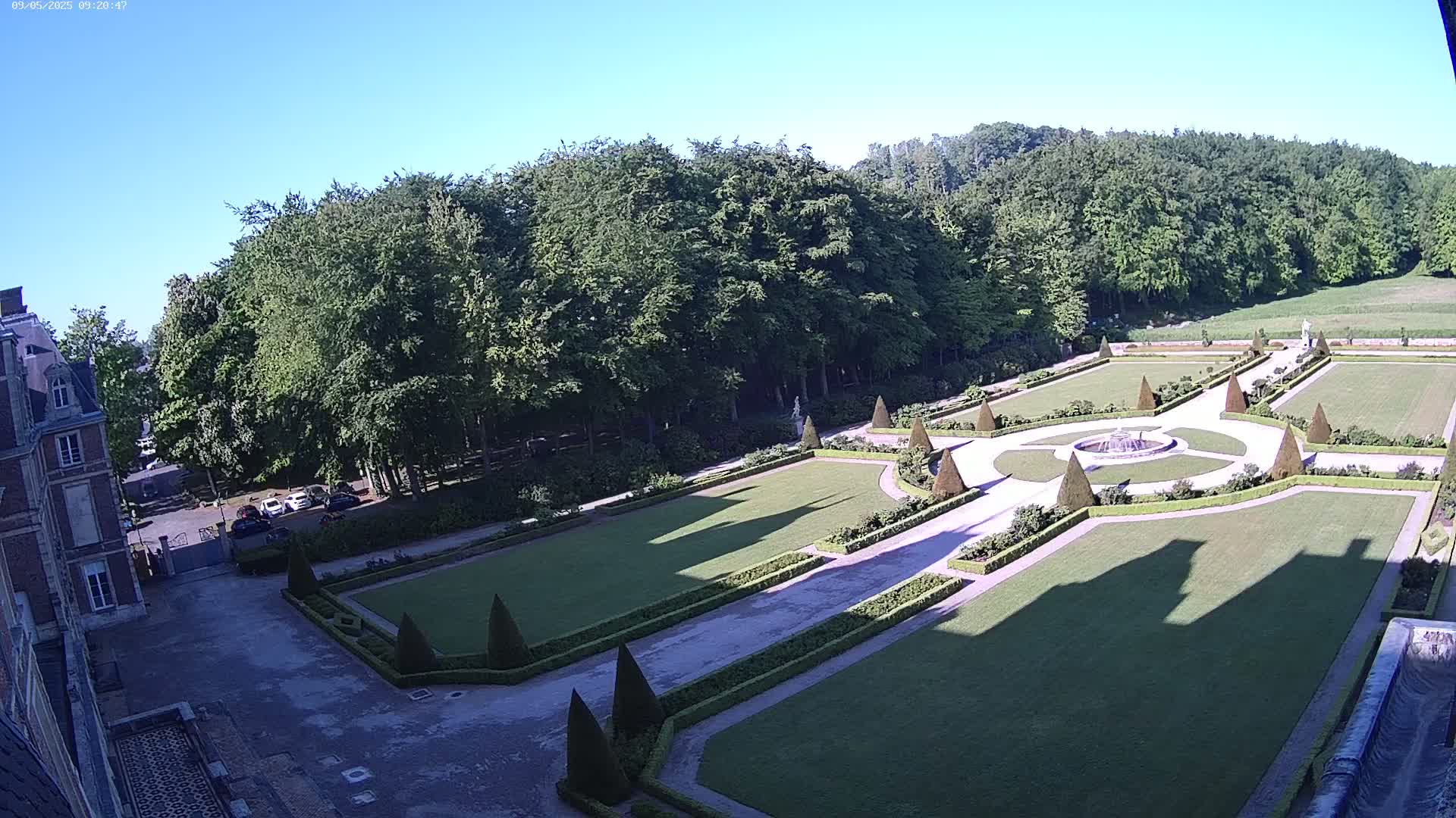 A formal garden with manicured lawns, hedges, and a central fountain is situated next to a building under a clear blue sky.