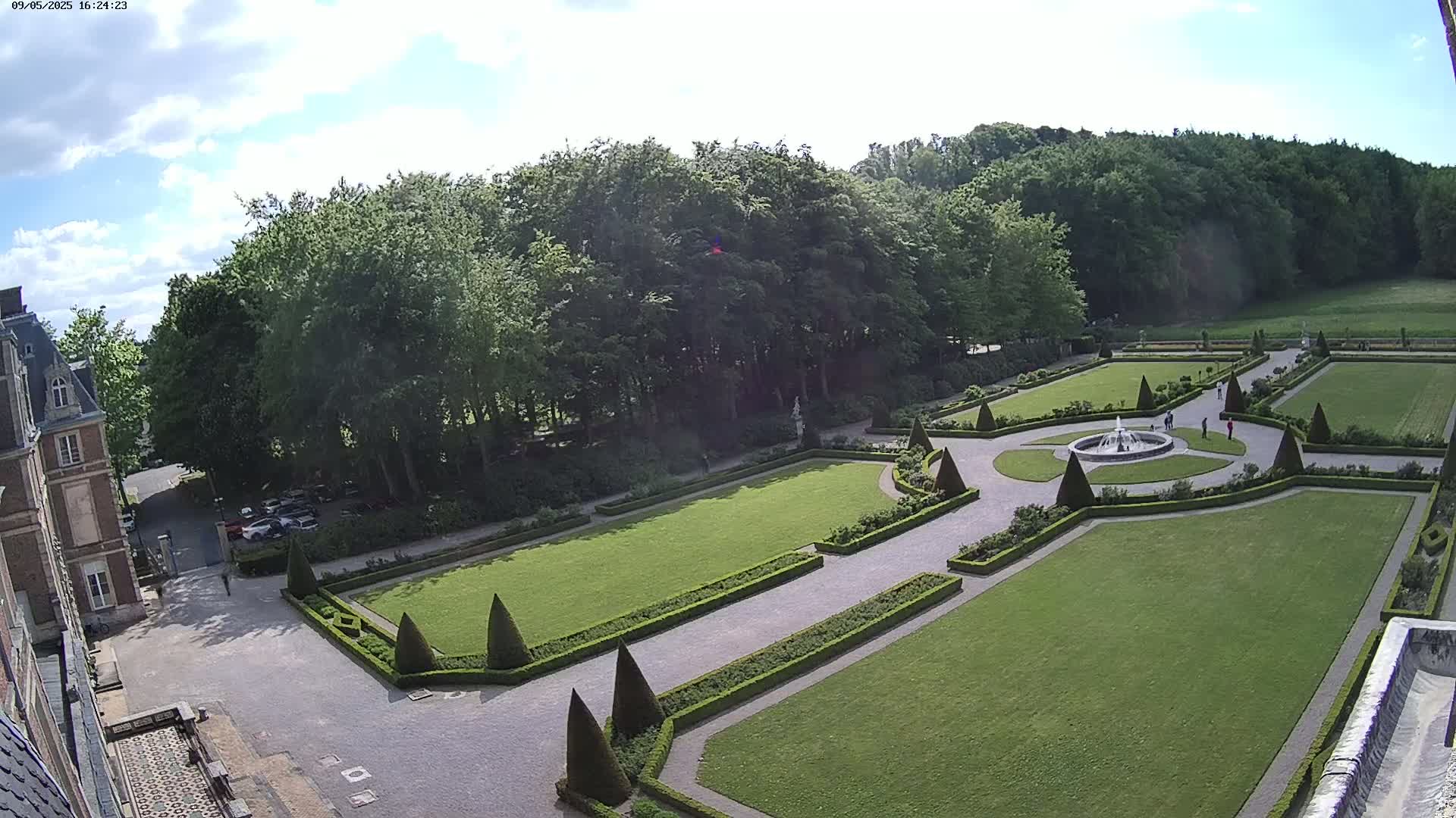 An aerial view shows a formal garden with a fountain, manicured lawns, and hedges, adjacent to a chateau under a partly cloudy sky.