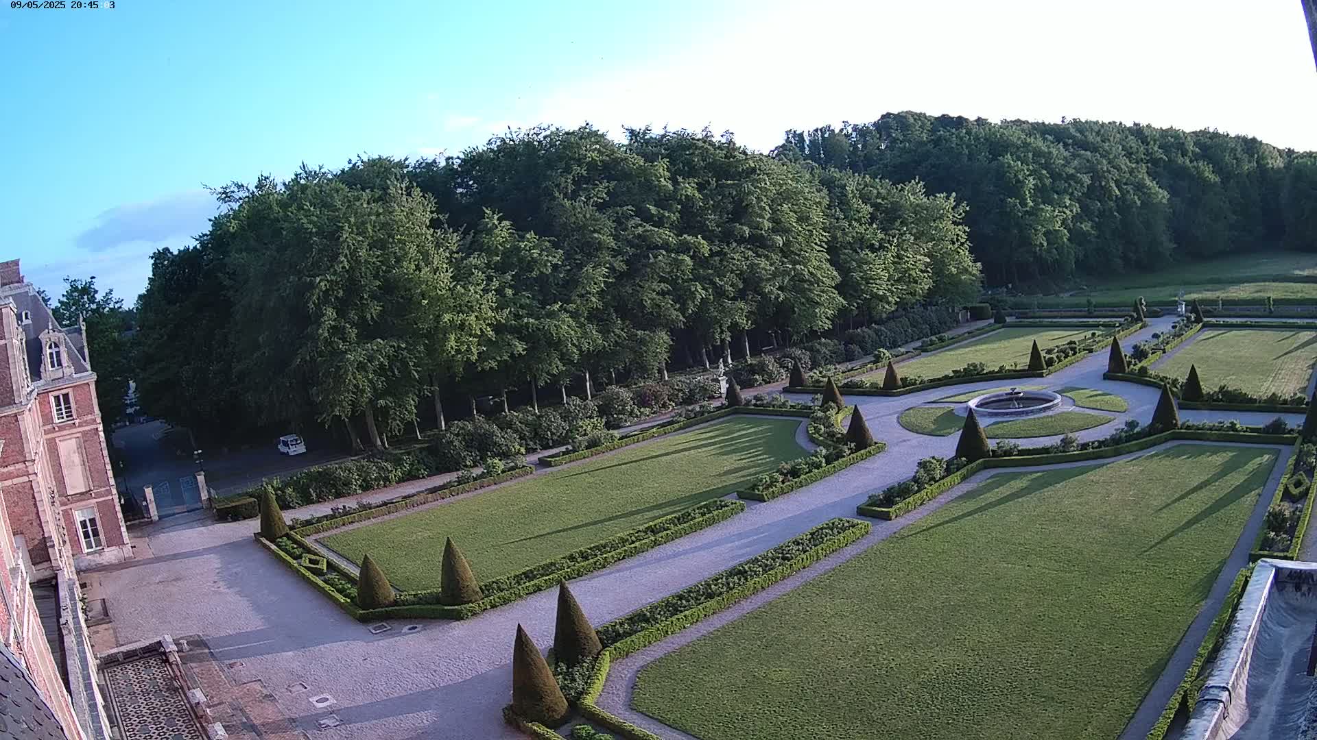 An aerial view shows a formal garden with manicured lawns, hedges, and a fountain, adjacent to a brick building under a clear blue sky.