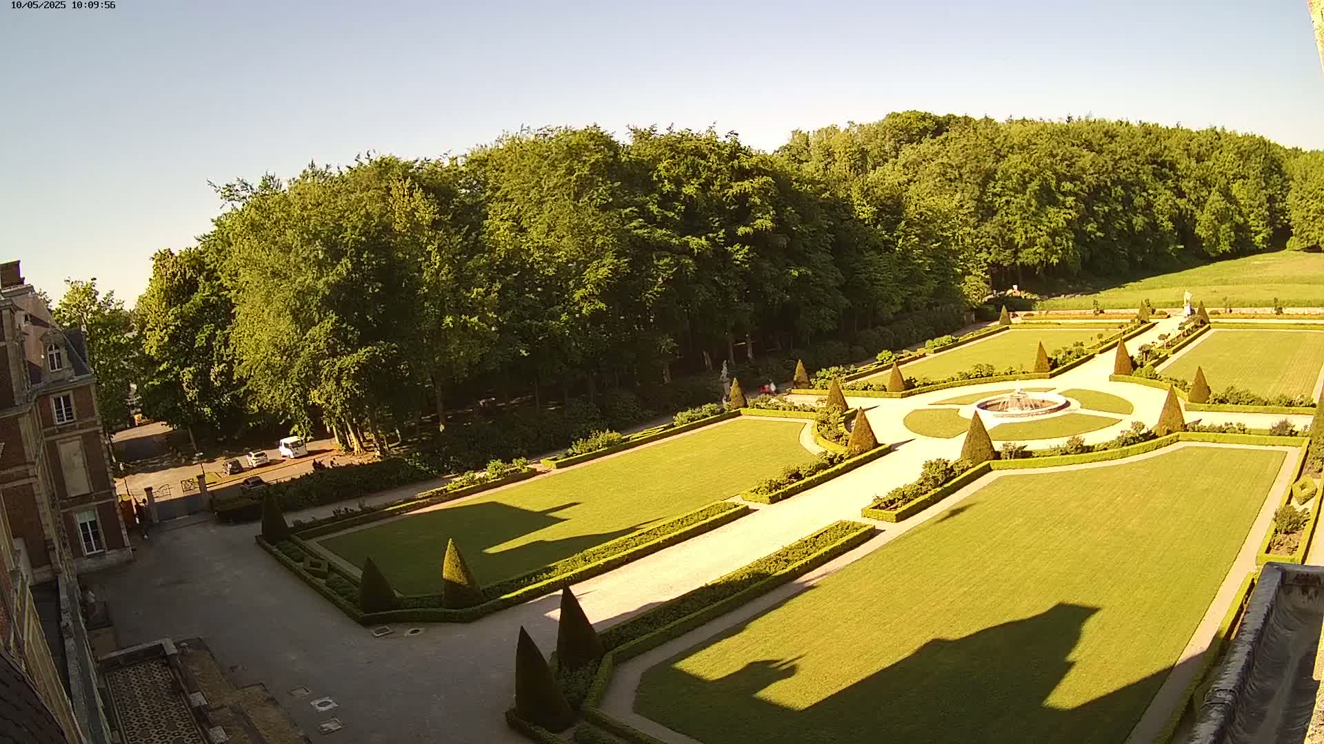 An aerial view shows a formal garden with manicured lawns, hedges, and a central fountain, bordered by a large forest under a clear, sunny sky.