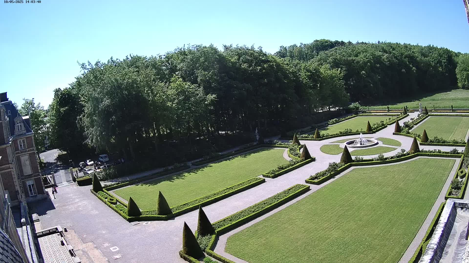 An aerial view shows a formal garden with manicured lawns, hedges, and a fountain, adjacent to a building and a wooded area under a clear, sunny sky.