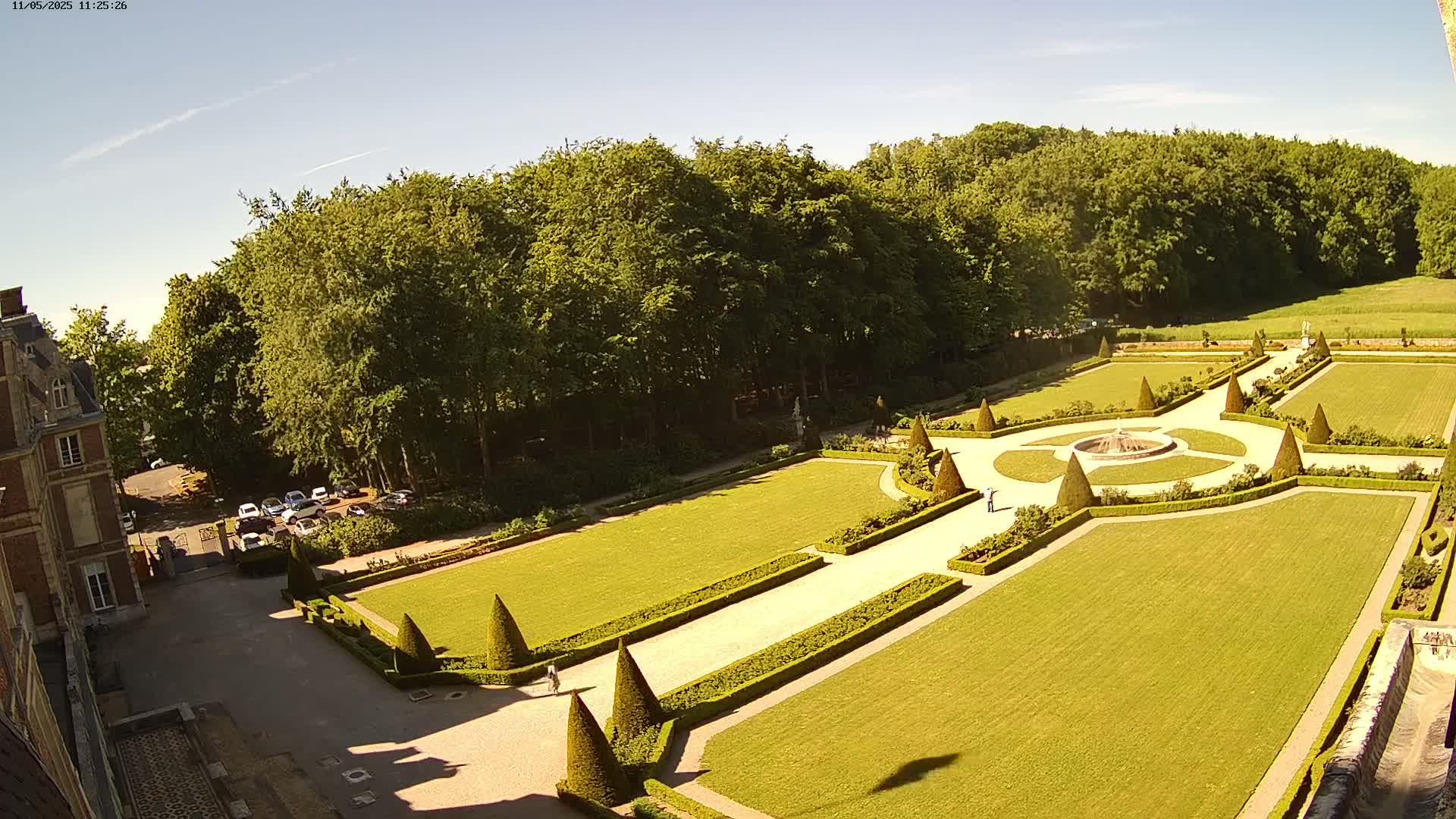 An aerial view shows a formal garden with manicured lawns, hedges, and a central fountain, adjacent to a building and a wooded area under a clear, sunny sky.