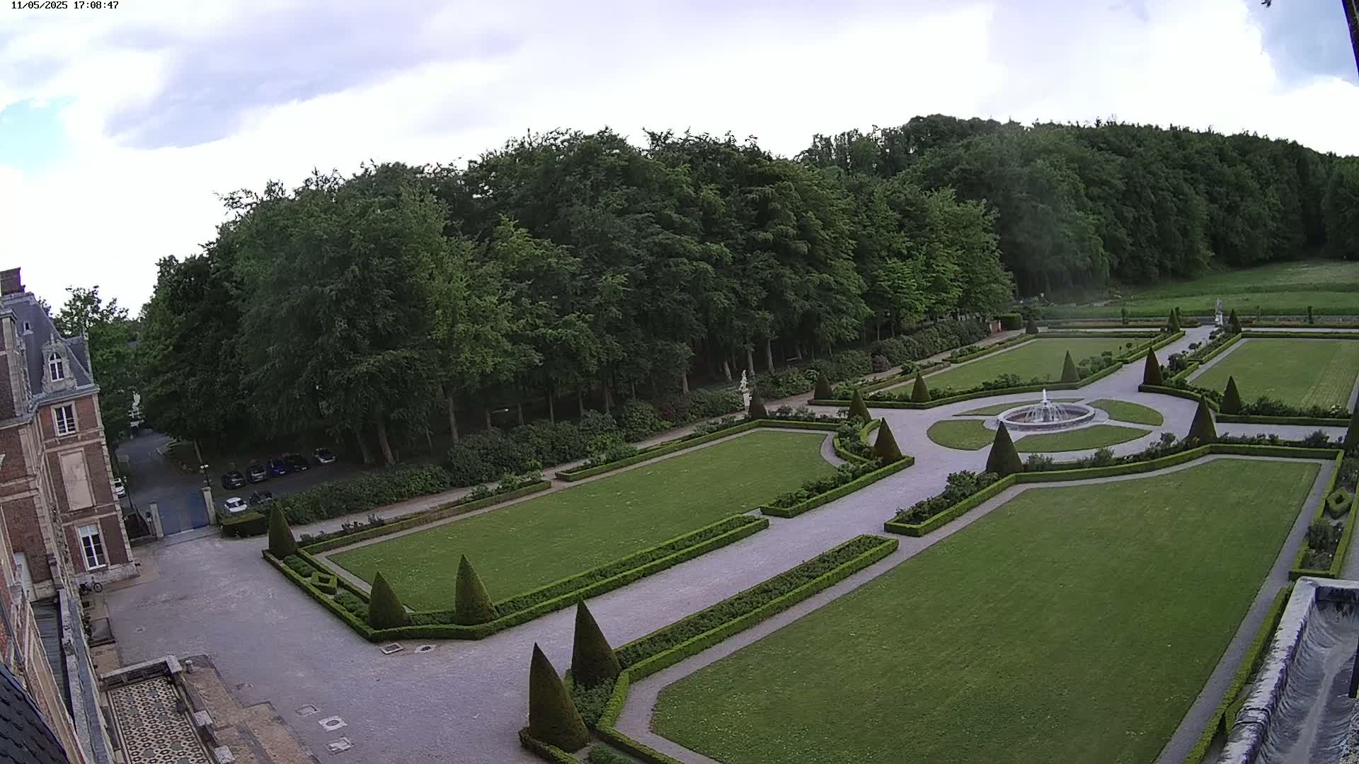 A formal garden with a fountain, manicured hedges, and green lawns is adjacent to a large building under a partly cloudy sky.