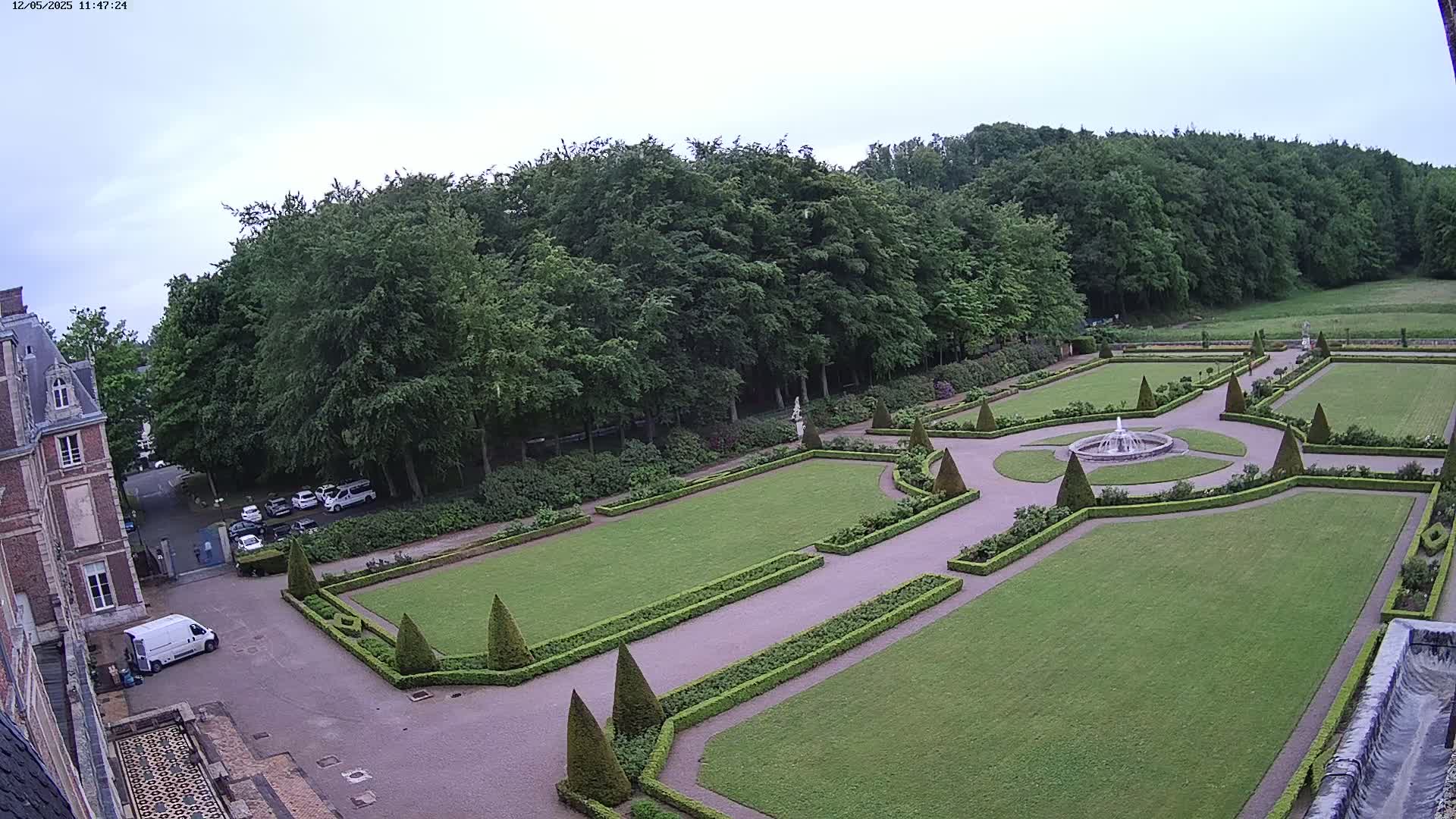 An aerial view shows a formal garden with a fountain, manicured lawns, and hedges, adjacent to a building and a wooded area under an overcast sky.