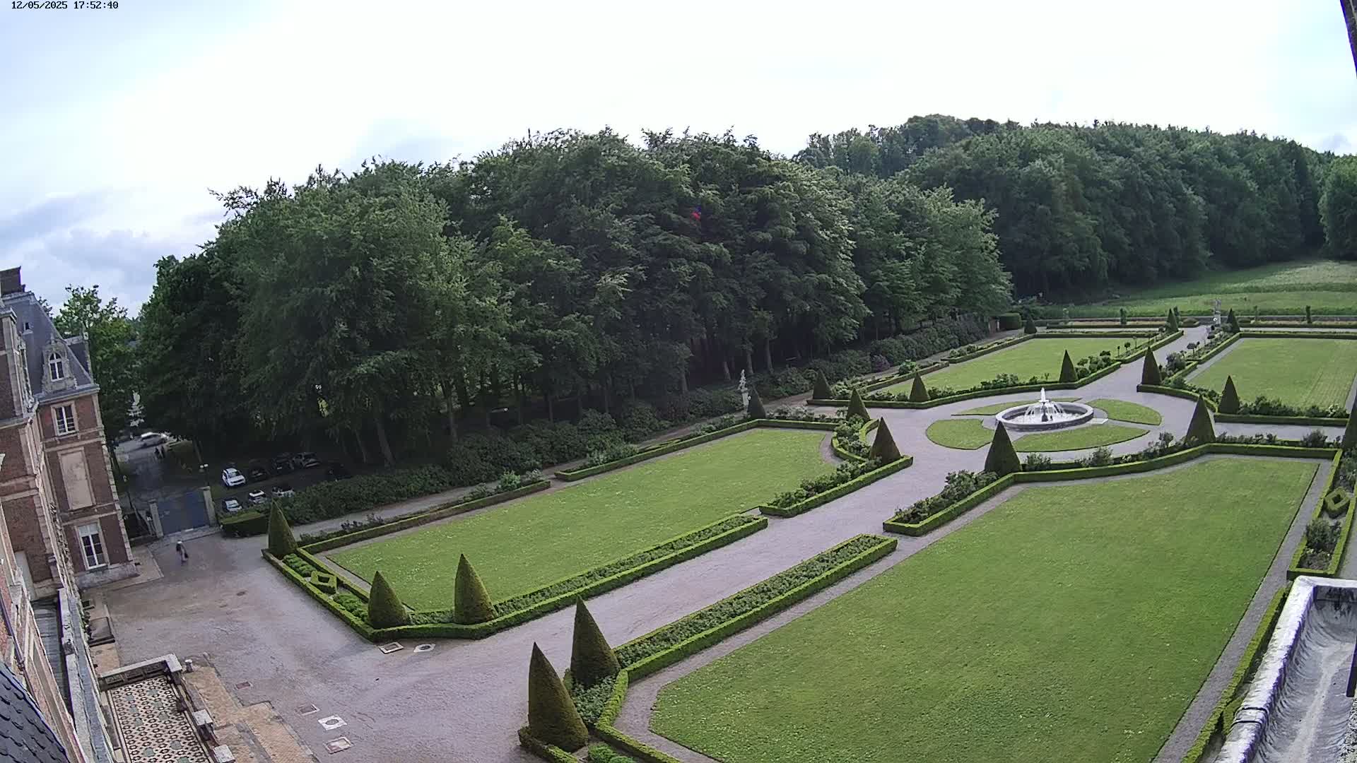 An aerial view shows a formal garden with a central fountain, manicured lawns, hedges, and a large building in the background under a partly cloudy sky.