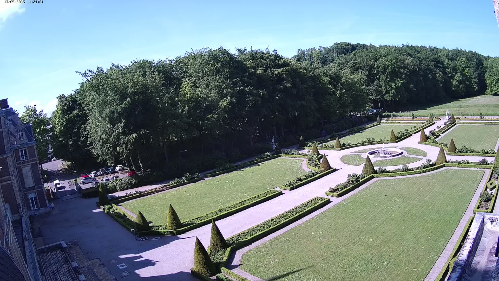 An elevated view shows a formal garden with manicured lawns, hedges, and a central fountain, adjacent to a large building under a clear blue sky.