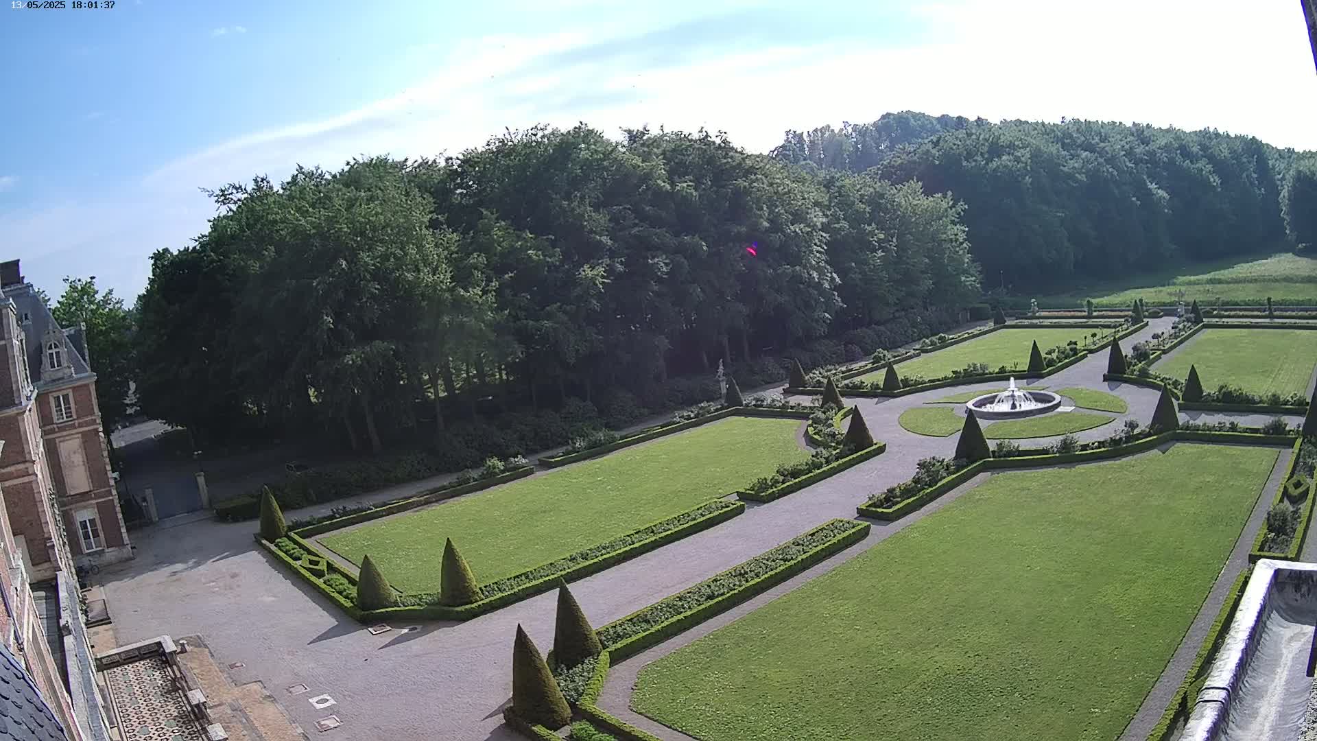 An expansive formal garden with manicured lawns, hedges, and a central fountain is situated behind a large brick building under a mostly sunny sky.