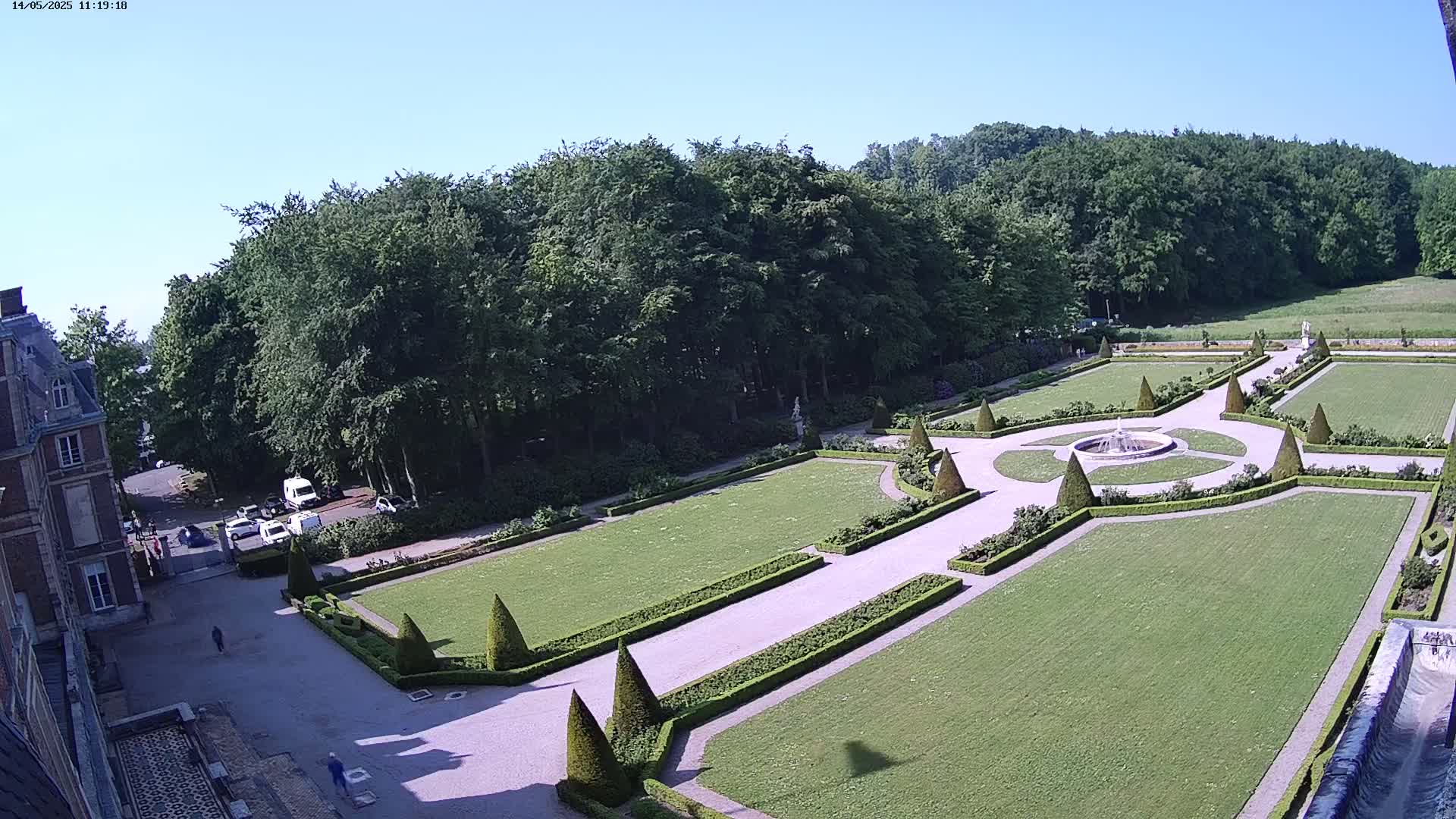 A high-angle view of a formal garden with manicured lawns, hedges, and a fountain, adjacent to a large building under a clear, sunny sky.