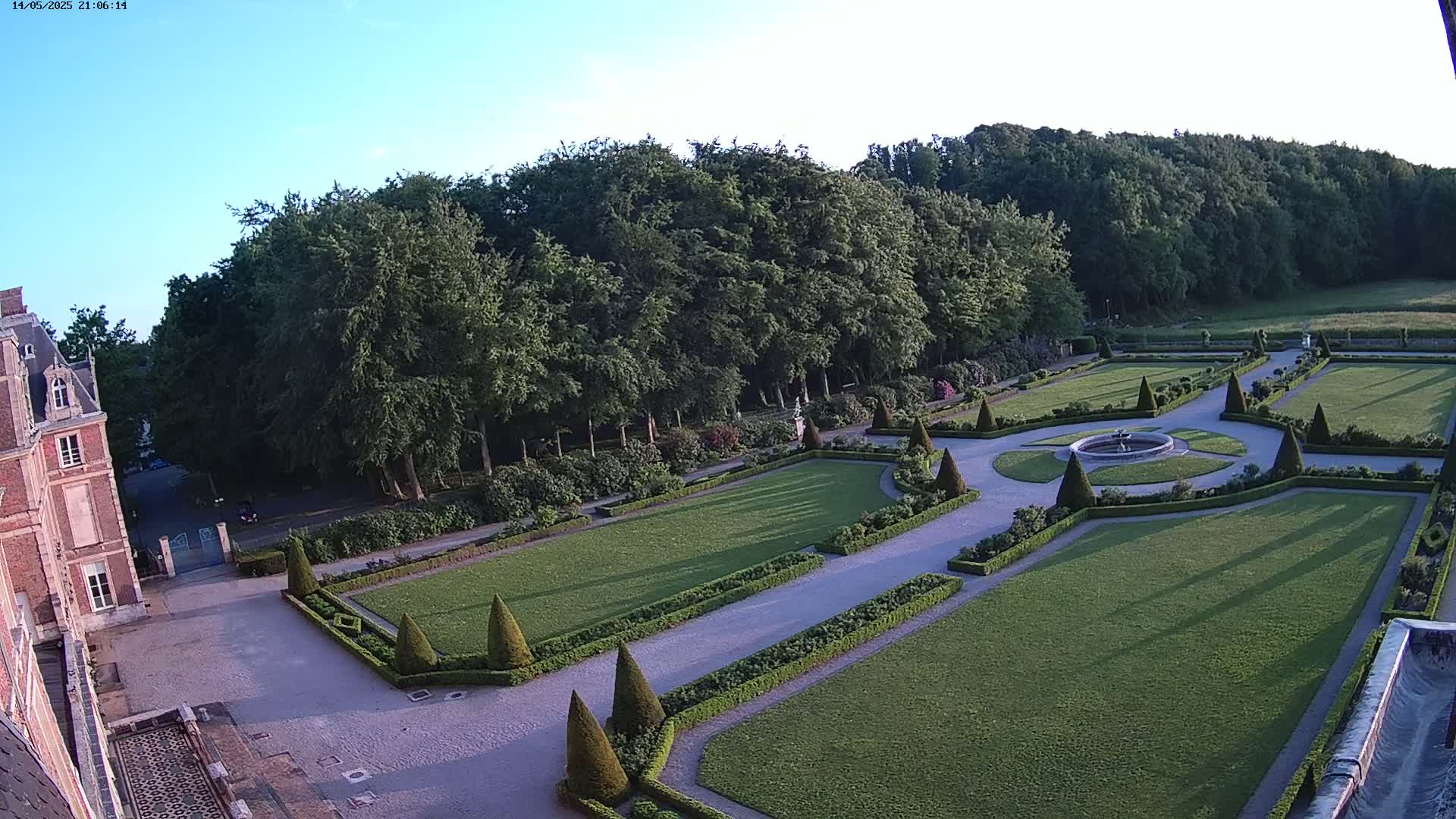 A formal garden with manicured lawns, hedges, and a central fountain is situated beside a large brick building under a clear, twilight sky.