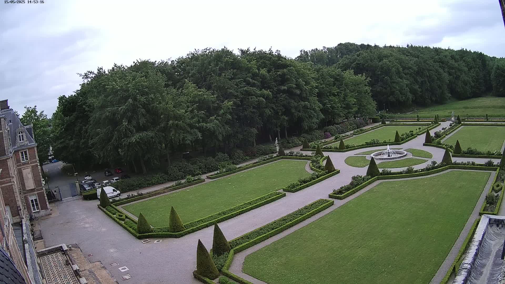 An aerial view shows a formal garden with manicured lawns, hedges, and a central fountain, adjacent to a large building under a mostly cloudy sky.
