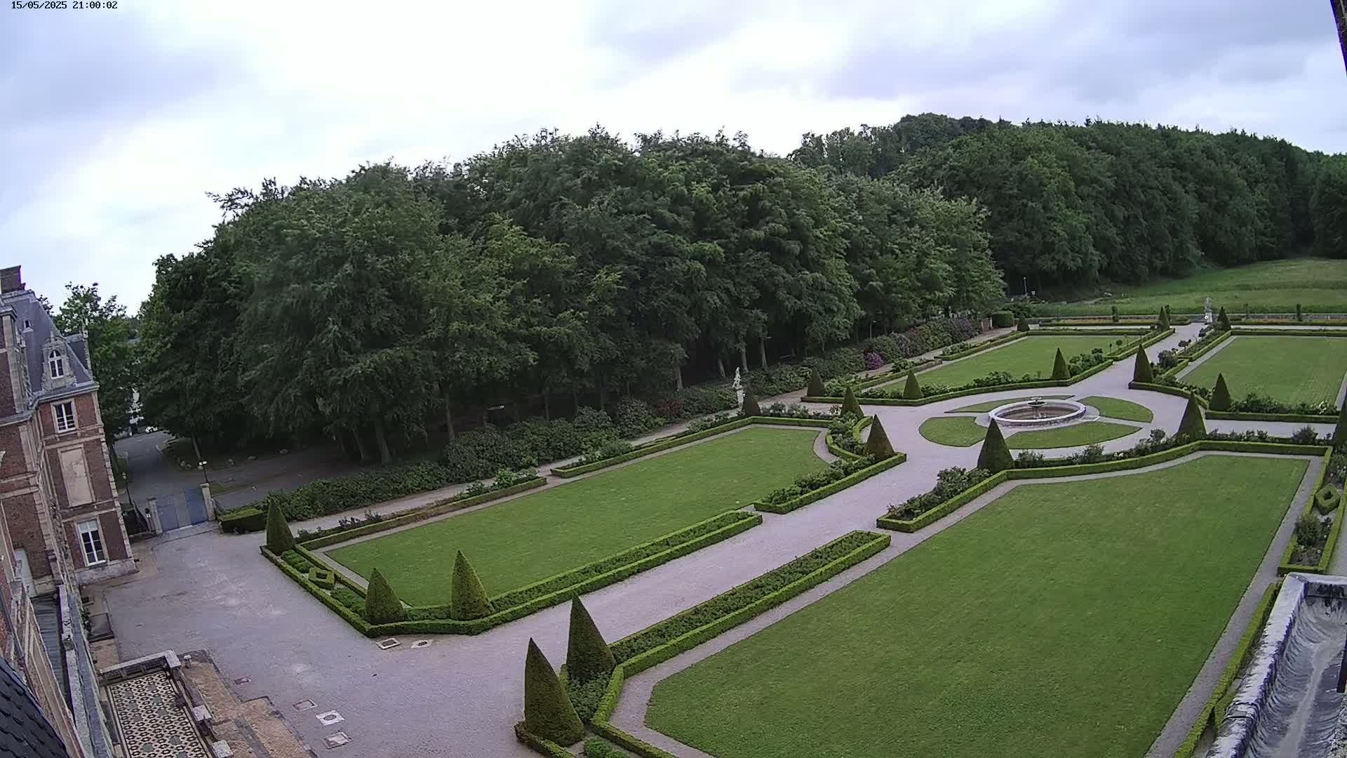 An overcast day reveals a formal garden with manicured lawns, hedges, and a central fountain, adjacent to a large brick building.
