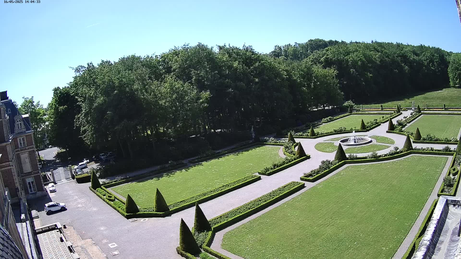 An aerial view shows a formal garden with manicured lawns, hedges, and a fountain, adjacent to a building and a wooded area under a clear blue sky.