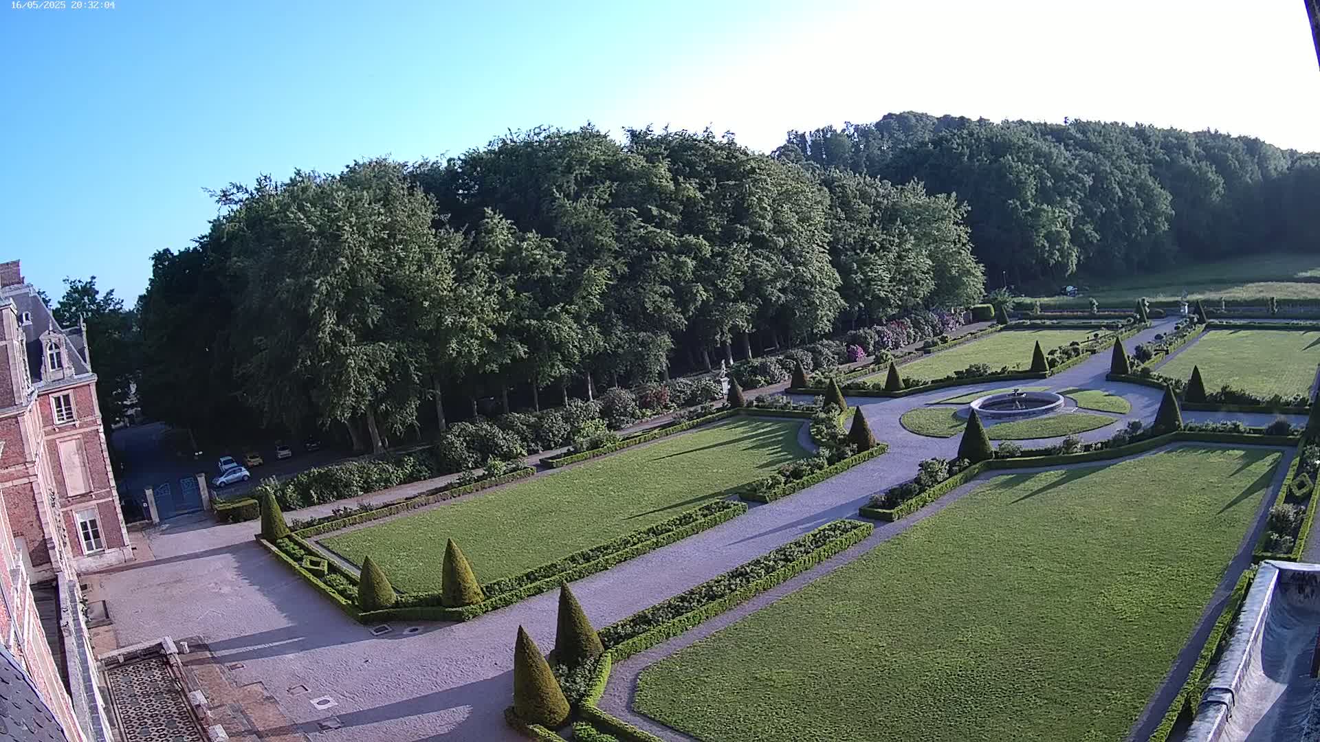 An expansive formal garden with manicured lawns, hedges, and a central fountain, bordered by a large forest and a brick building on a clear day.