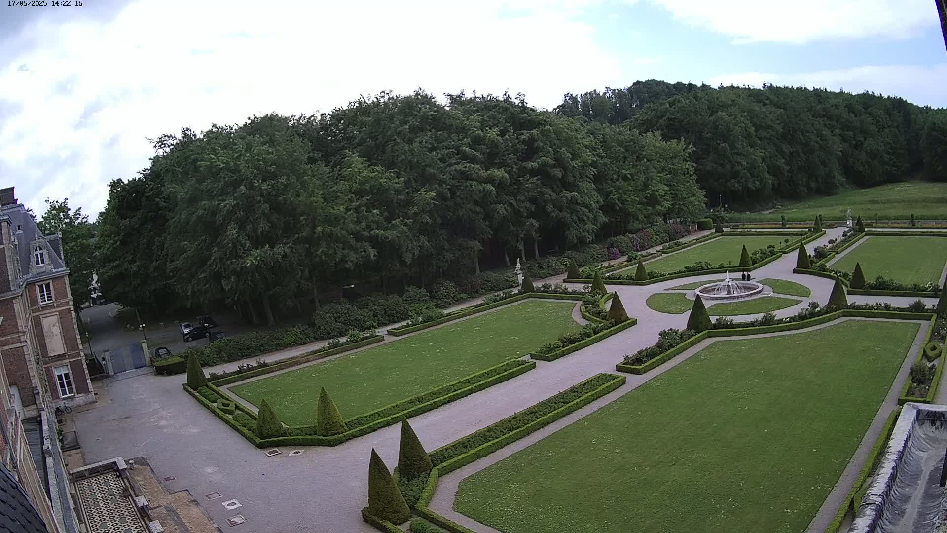 An aerial view shows a formal garden with manicured lawns, hedges, and a central fountain, adjacent to a large brick building under a partly cloudy sky.