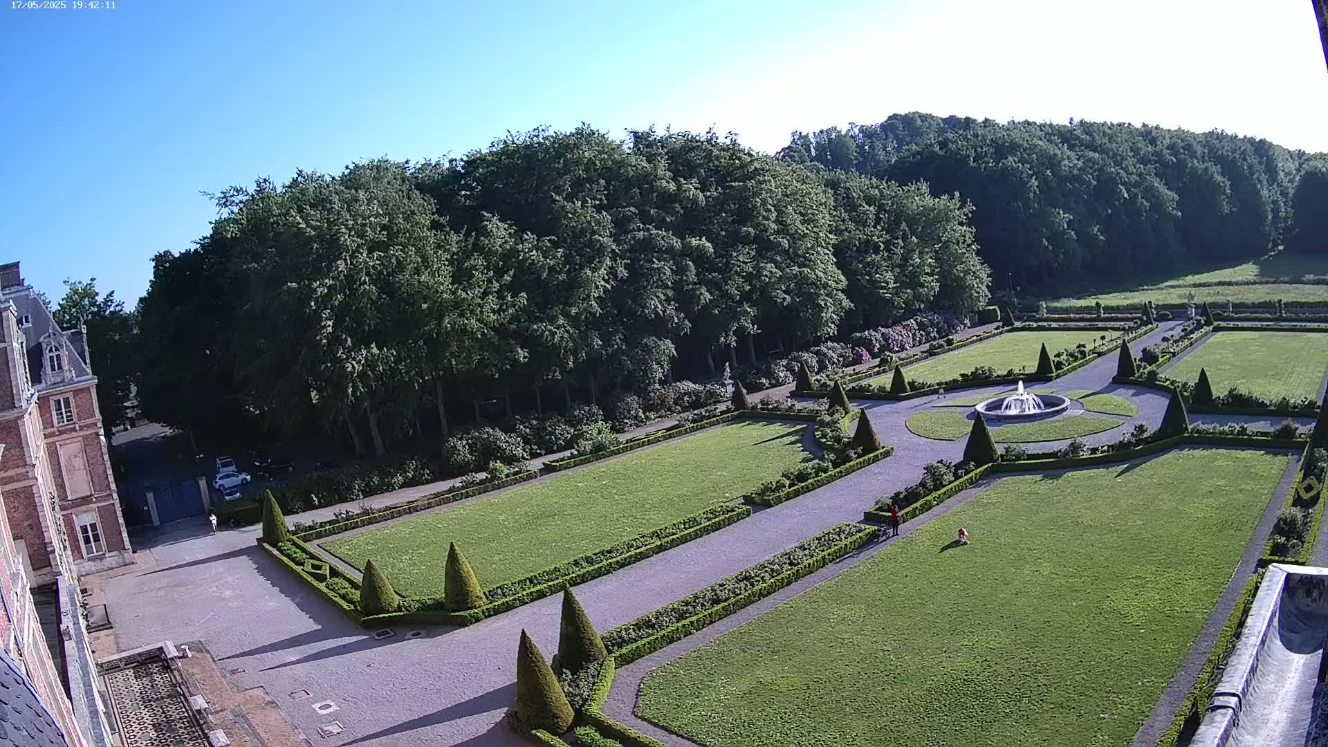 An elevated view shows a formal garden with manicured lawns, hedges, and a fountain, adjacent to a large building under a clear blue sky.