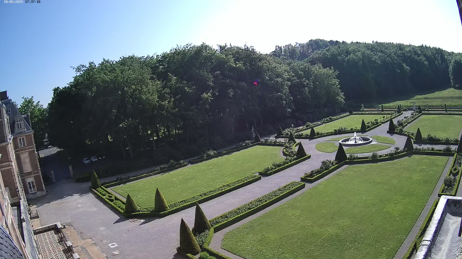 An elevated view shows a formal garden with manicured lawns, hedges, and a central fountain, adjacent to a large building under a clear, sunny sky.