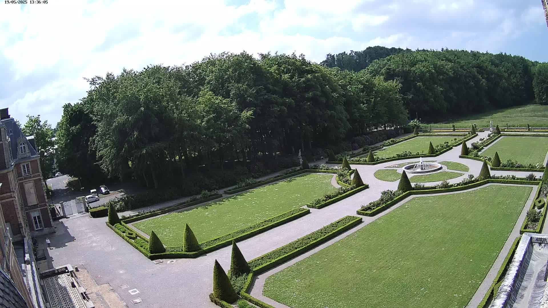 An aerial view shows a formal garden with manicured lawns, hedges, and a central fountain, adjacent to a large building under a partly cloudy sky.