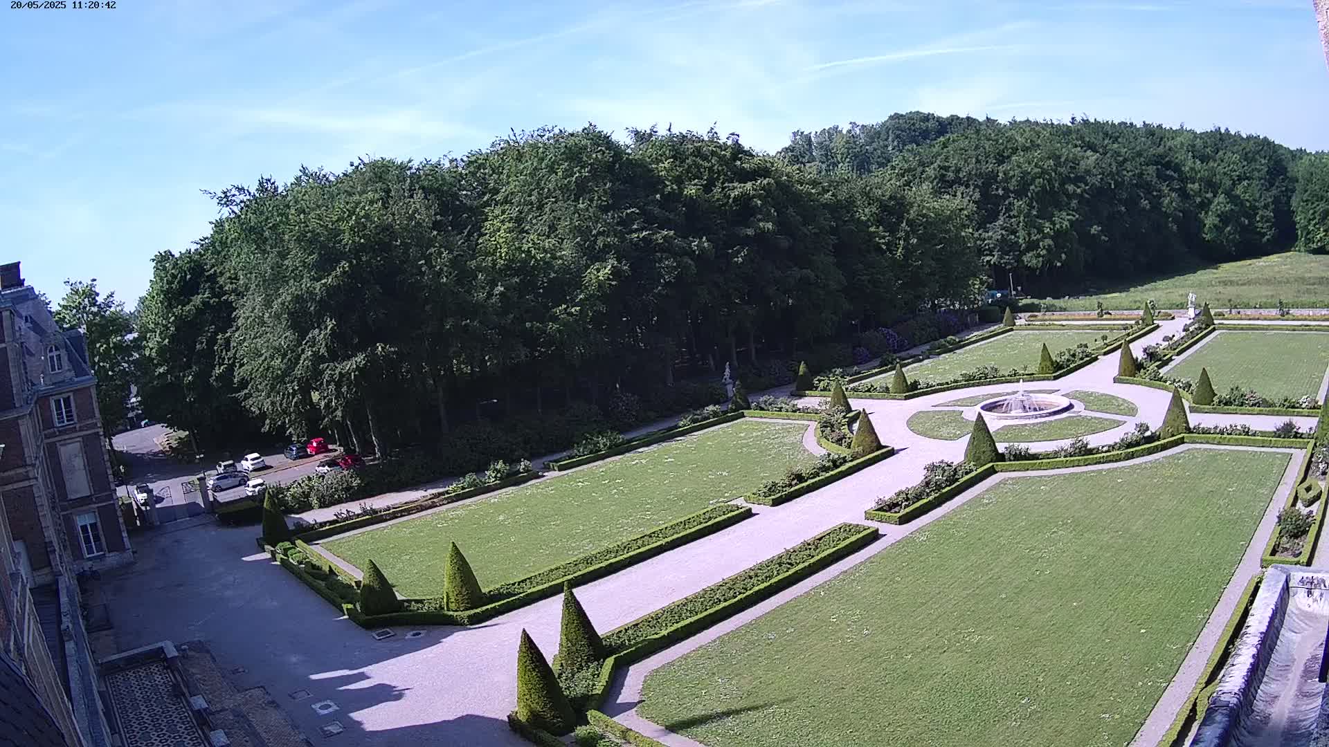 An aerial view shows a formal garden with manicured lawns, hedges, and a central fountain, adjacent to a large building and a wooded area under a clear, sunny sky.
