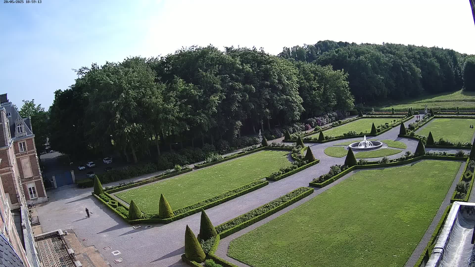 A formal garden with a fountain is situated beside a large building, all surrounded by a dense forest under a clear sky.
