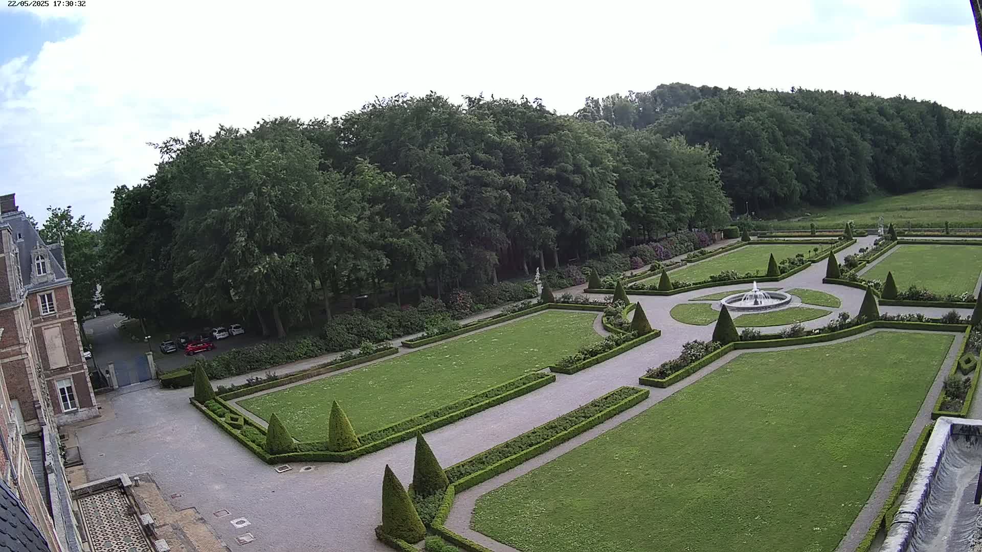 An aerial view shows a formal garden with manicured lawns, hedges, and a central fountain, adjacent to a large building under a partly cloudy sky.