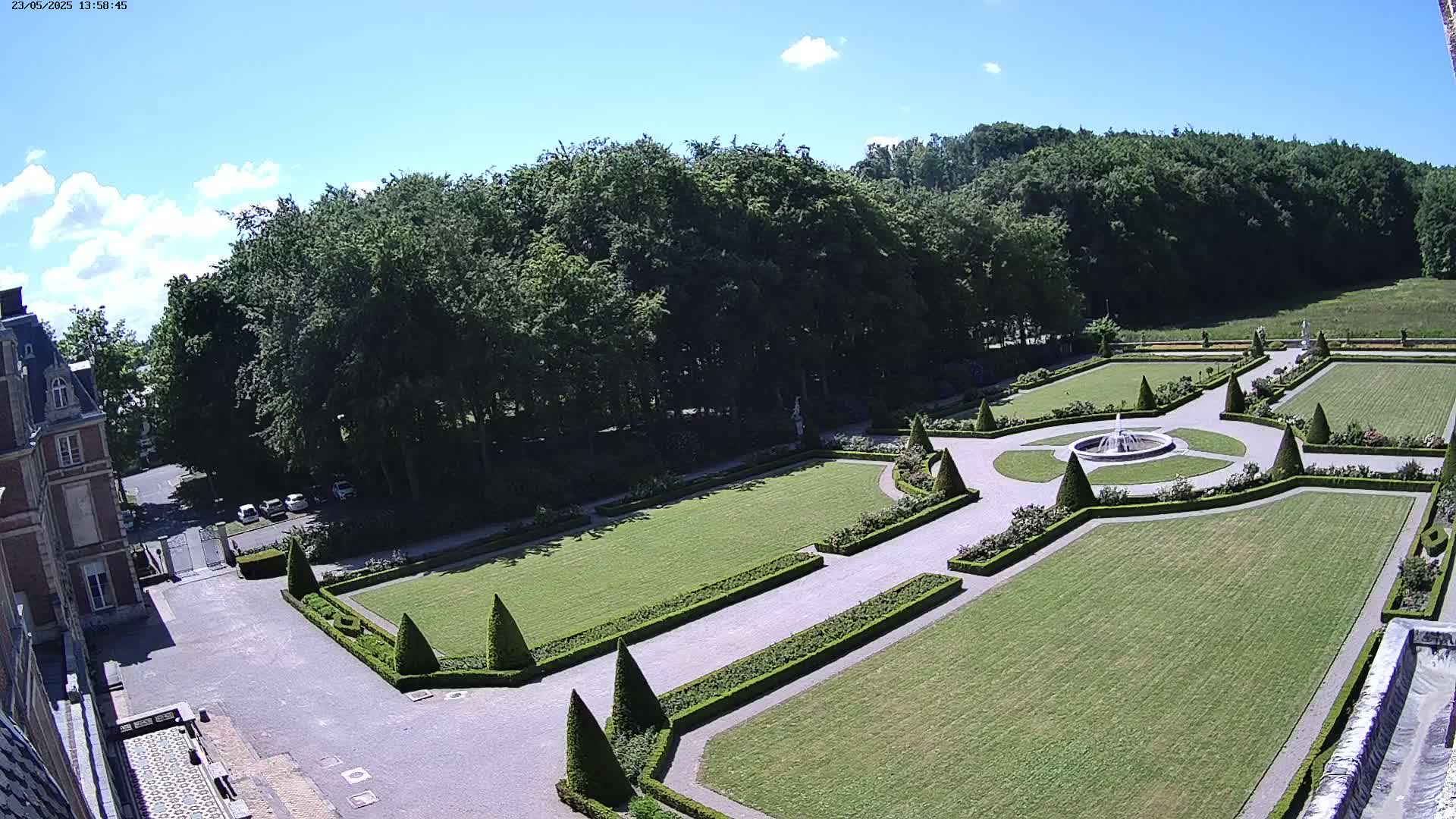 An aerial view shows a formal garden with manicured lawns, hedges, and a fountain, adjacent to a large building under a mostly sunny sky.